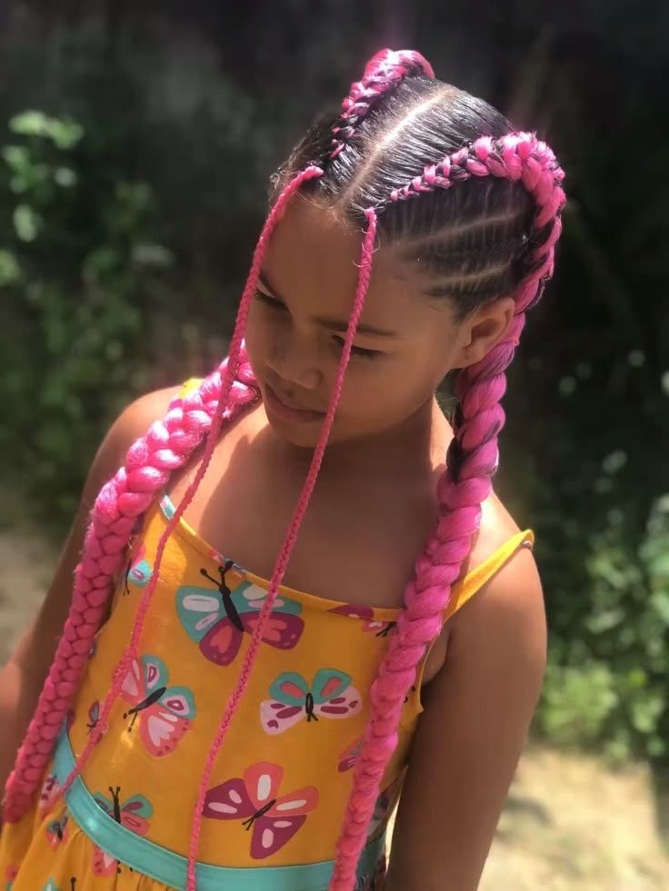 Niña pequeña con cabello rosa trenzado y vestido amarillo con mariposas, de pie al aire libre bajo la luz del sol.