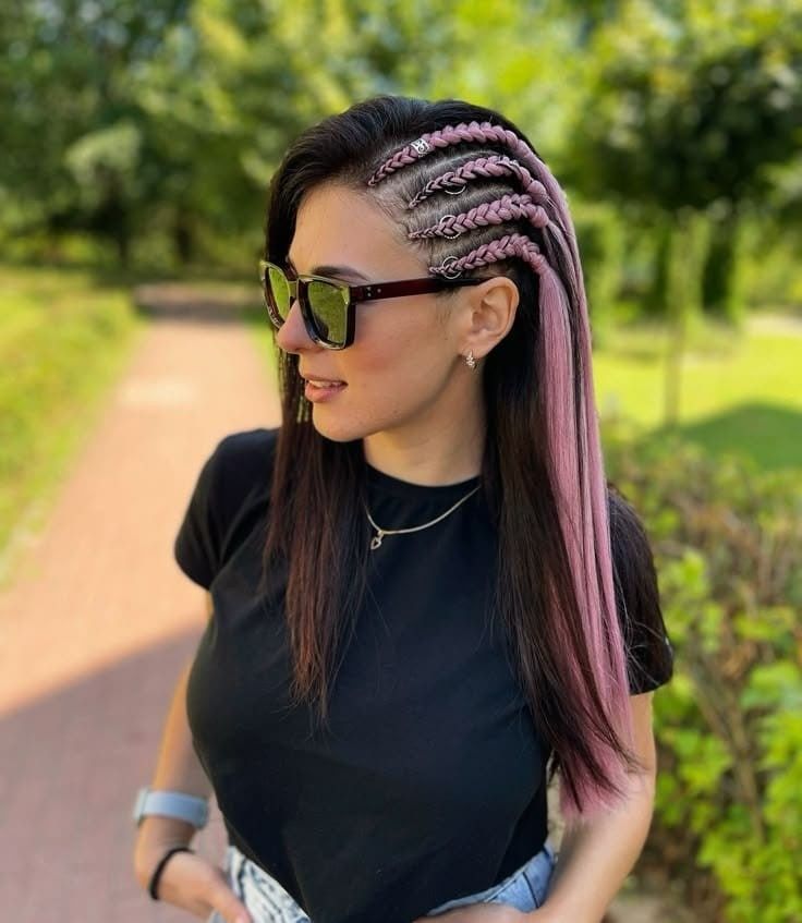 Mujer con largas trenzas rosas y gafas de sol de pie en un sendero soleado en un parque.