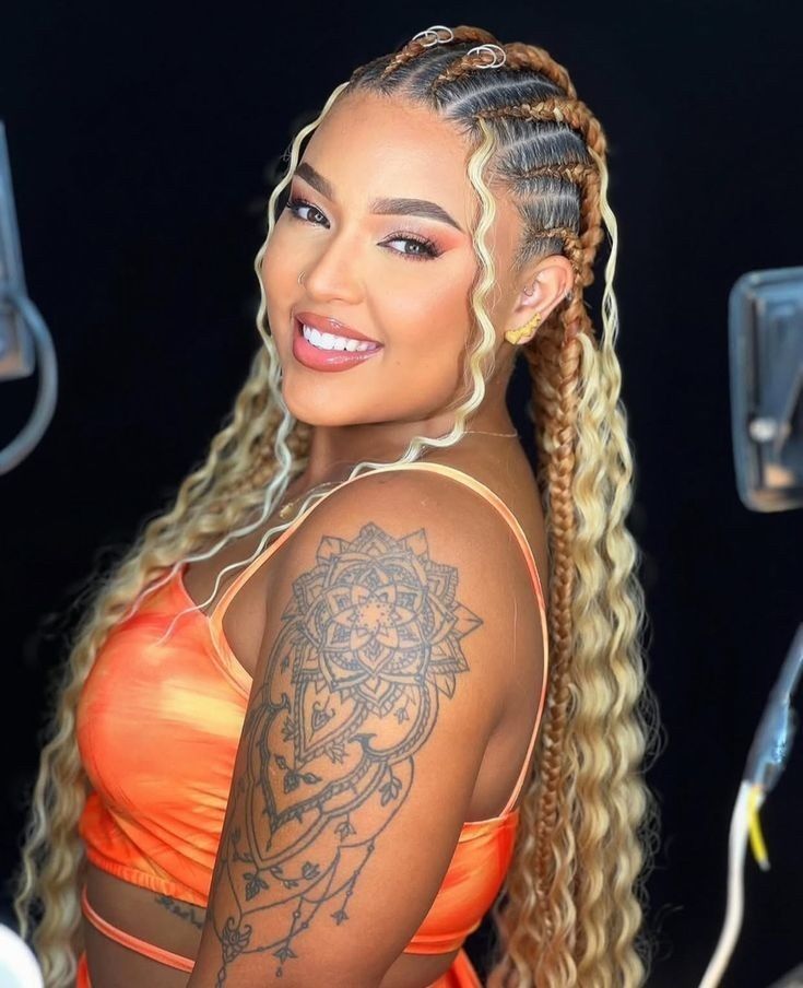 Mujer sonriente con cabello trenzado y blusa naranja, posando contra un fondo oscuro, mostrando un brazo tatuado.