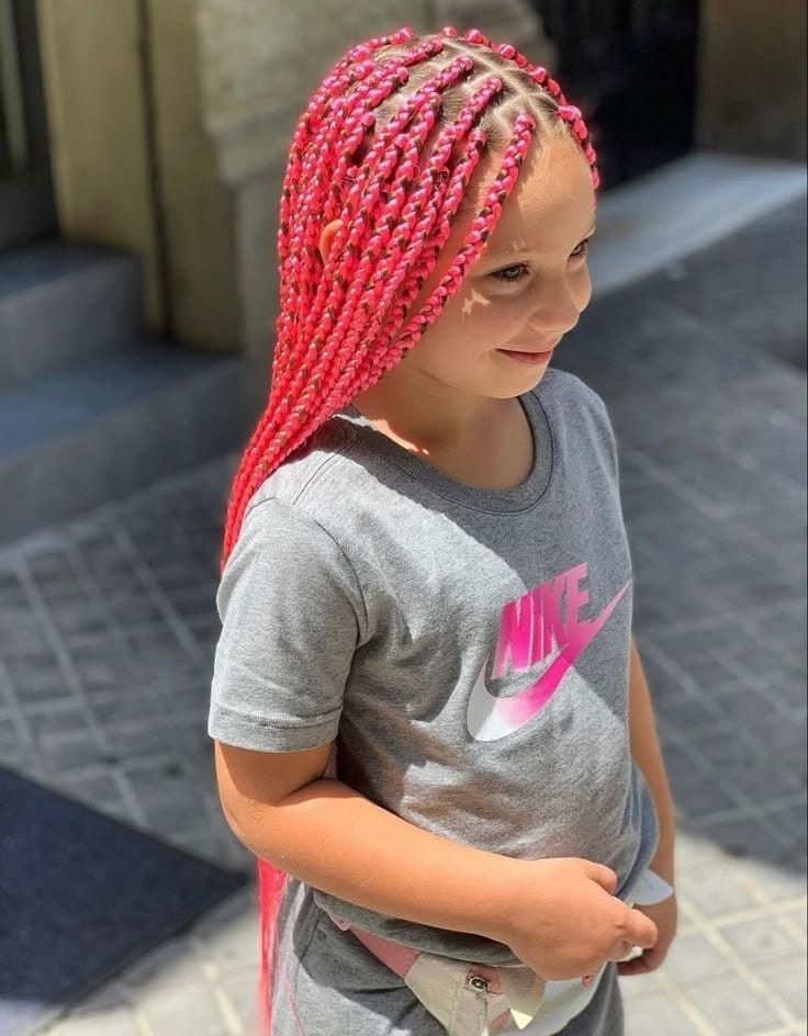 Niña con el pelo trenzado de color rosa brillante y una camiseta gris de Nike de pie al aire libre bajo la luz del sol.