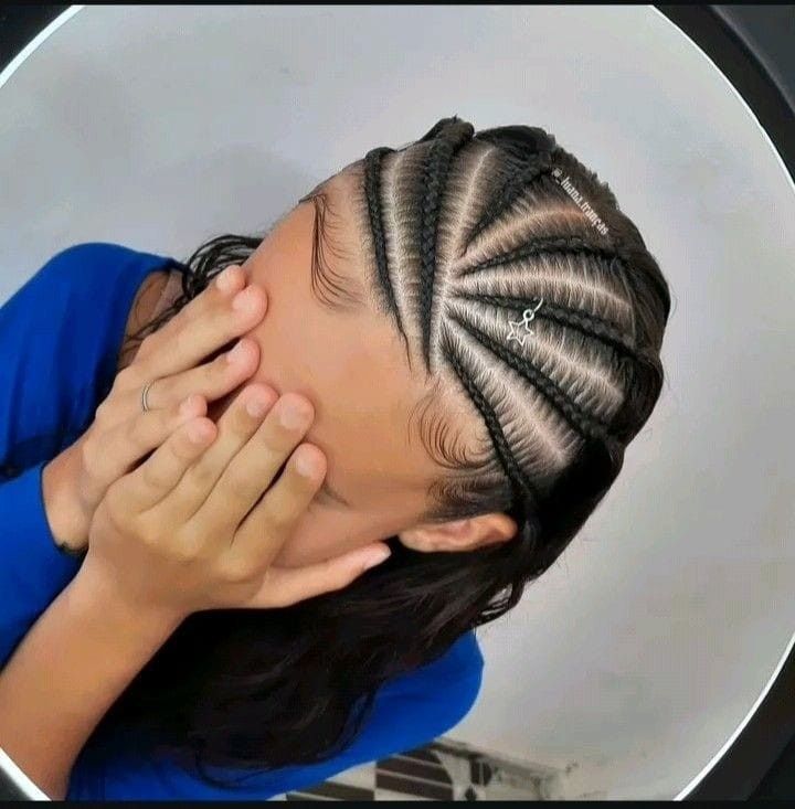 Persona con trenzas africanas que le cubren la cara con ambas manos, vestida con una camisa azul sobre un fondo gris.