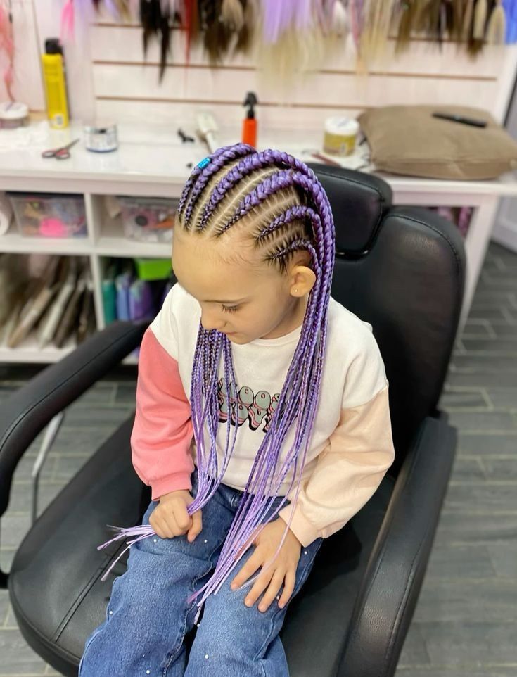Niña con largas trenzas moradas sentada en una silla de peluquería, mirando hacia abajo, vestida con una camiseta rosa