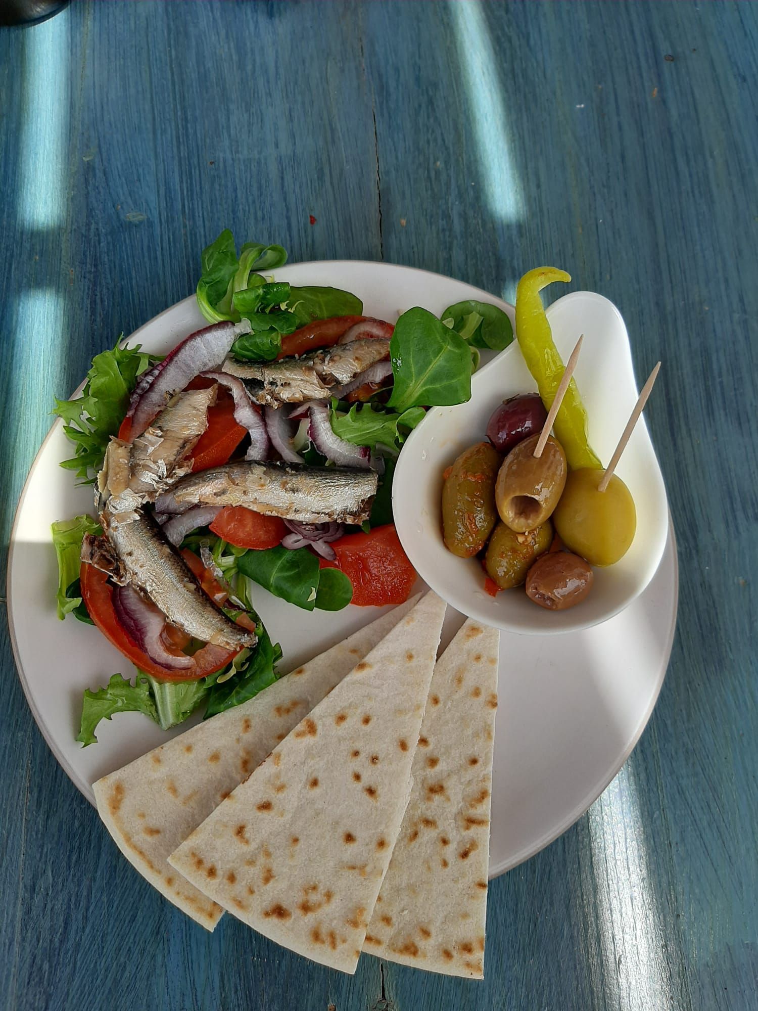 Un plato blanco cubierto con ensalada, pan pita y aceitunas sobre una mesa azul.