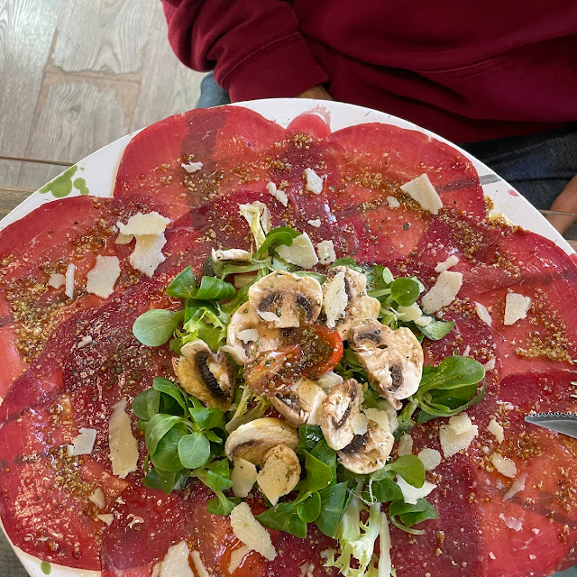 Carpaccio con champiñones, parmesano y verduras en un plato, con rodajas de carne roja visibles.