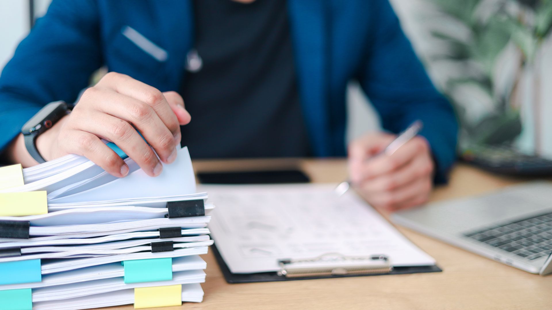 Des mains trient des piles de papiers avec des post-it colorés à côté d'un bloc-notes et d'un ordinateur portable.