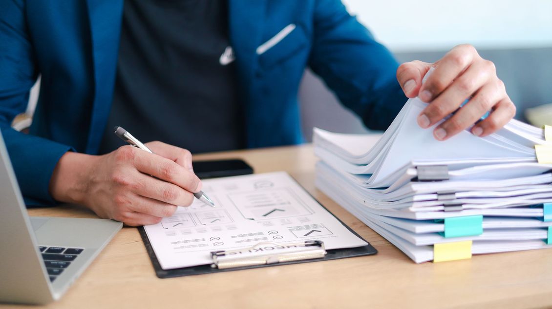 Une personne en chemise bleue écrit sur une liste de contrôle à côté d'une pile de documents posés sur un bureau.