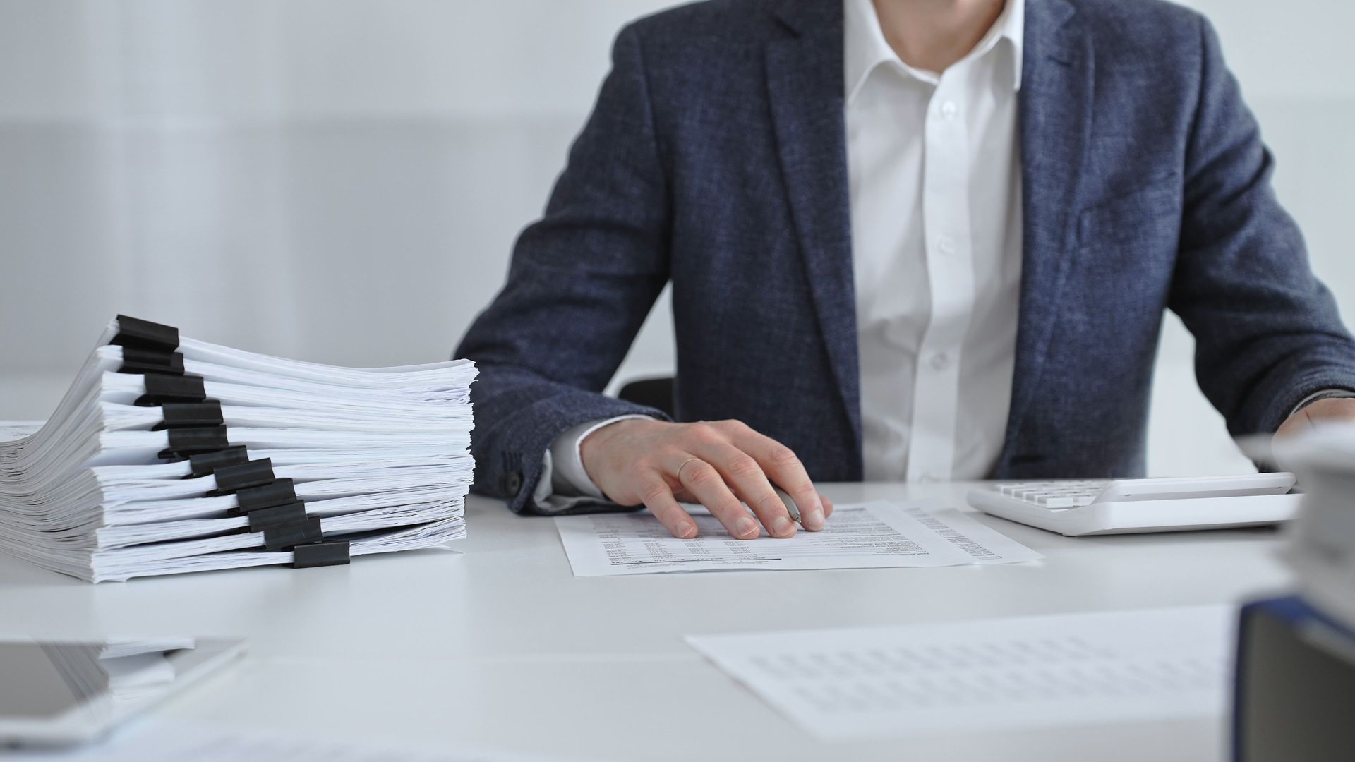 Une personne vêtue d'un costume est assise à un bureau, entourée de piles de documents.