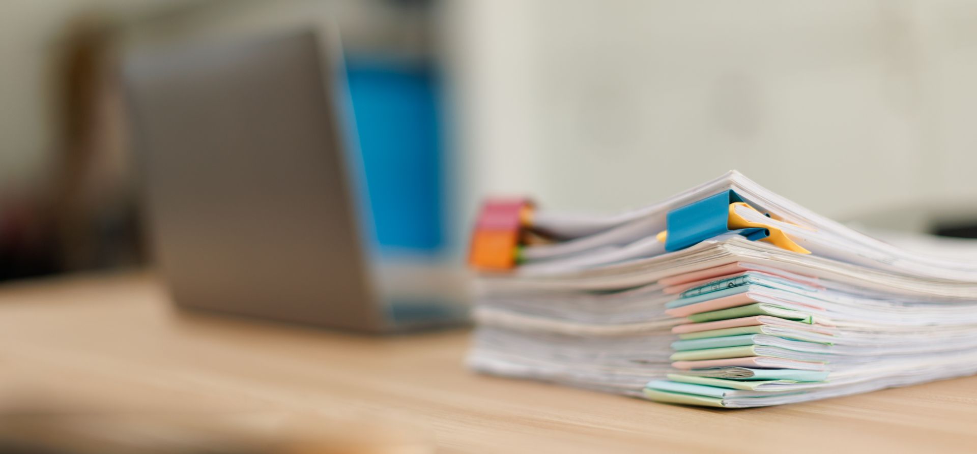Pile de papiers sur un bureau à côté d'un ordinateur portable, avec des post-it colorés.