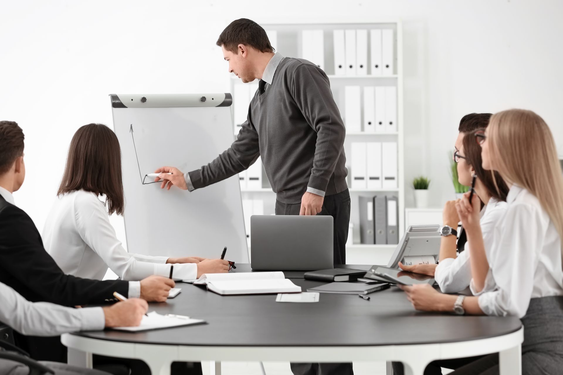 Réunion d'affaires dans un bureau lumineux, avec un homme présentant un graphique au tableau blanc.