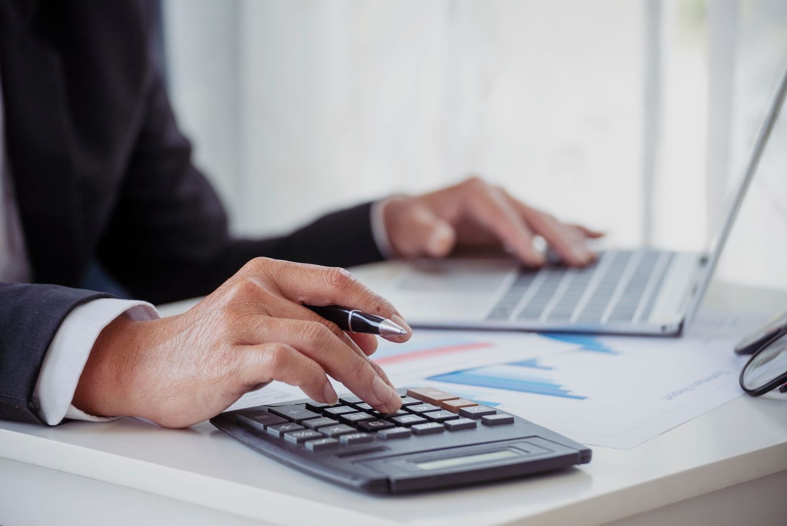 Des mains tapant sur calculatrice avec un ordinateur portable sur un bureau où figurent des graphiques financiers.