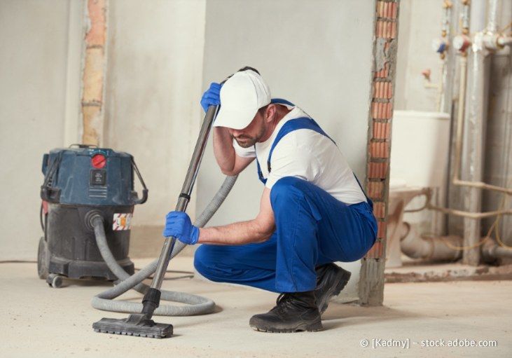 Ein Mann in blauem Overall reinigt mit einem Staubsauger den Boden einer Baustelle.