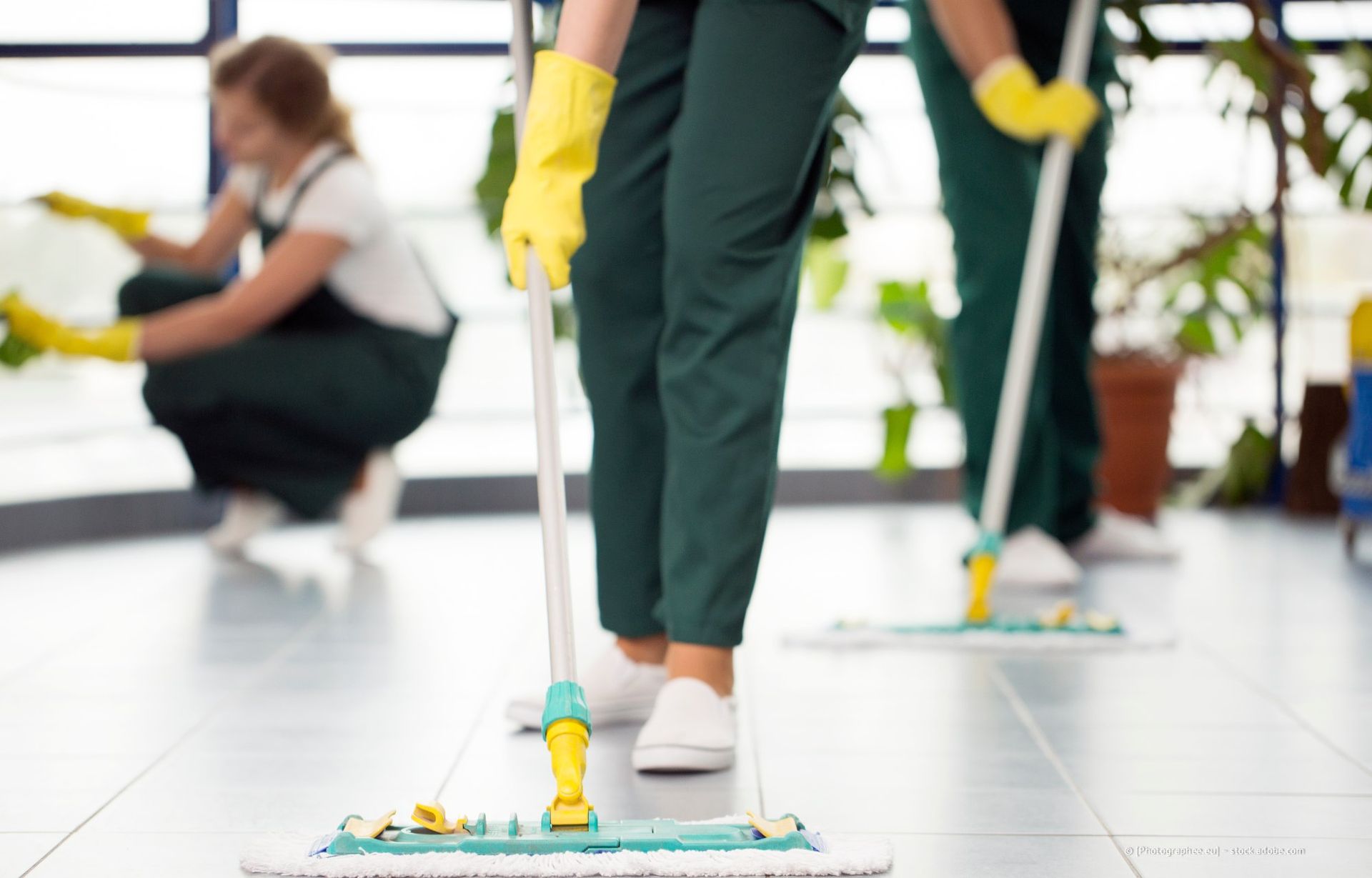 Drei Personen in grünen Hosen und gelben Handschuhen wischen einen Boden.