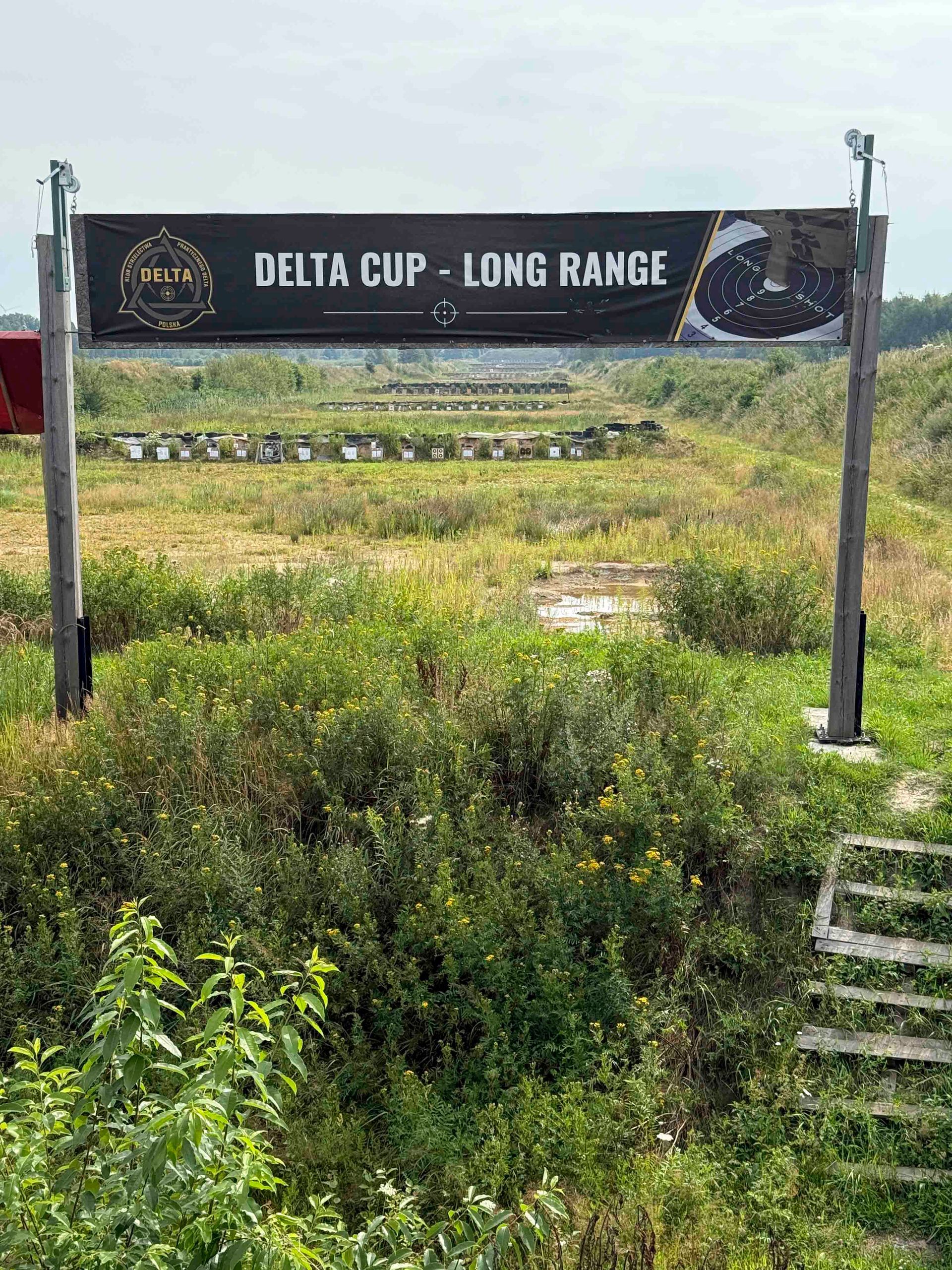 Hinweisschild für den Delta Cup Weitdistanzschießwettbewerb über einem grasbewachsenen Feld mit Zielstationen.