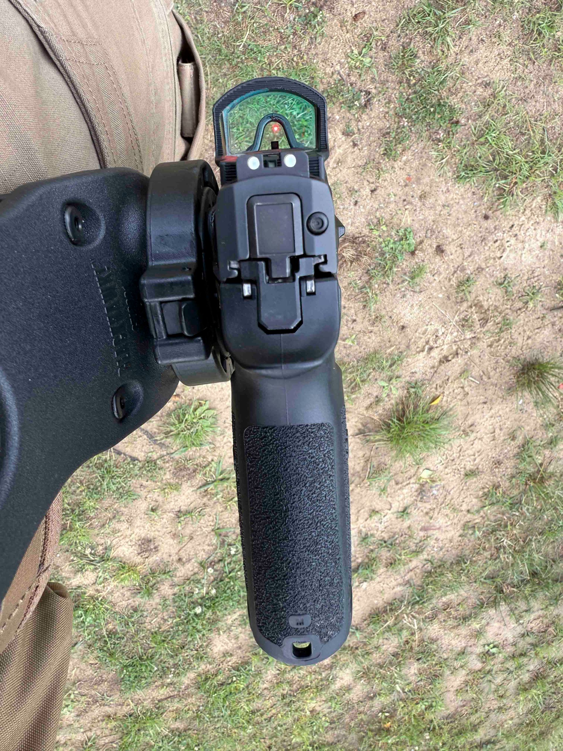 Schwarze Pistole mit Rotpunktvisier im Holster vor grasbewachsenem Hintergrund.