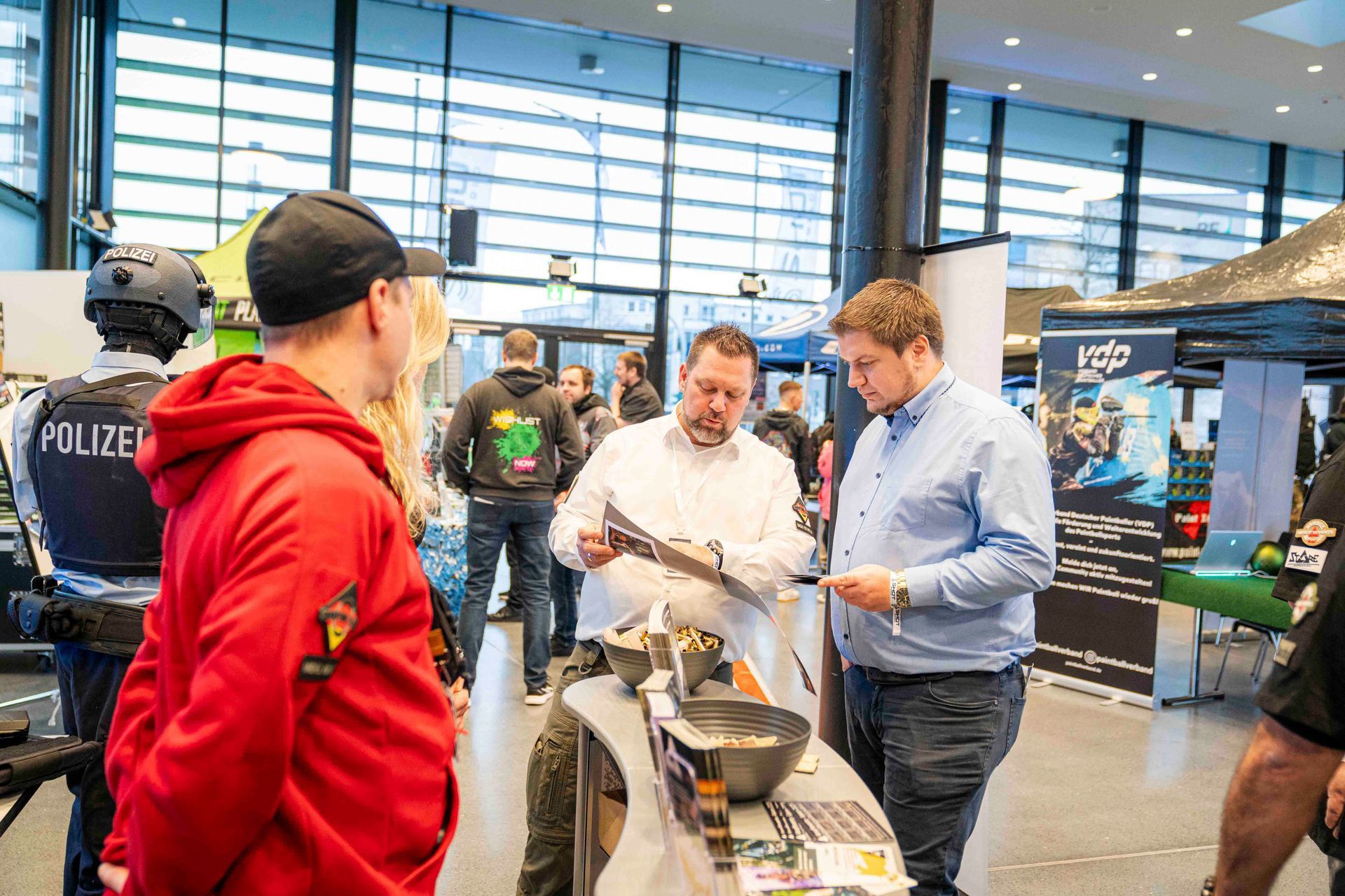 Menschen auf einer Messe; ein Mann betrachtet einen Gegenstand auf einem Tisch, zwei andere schauen zu.