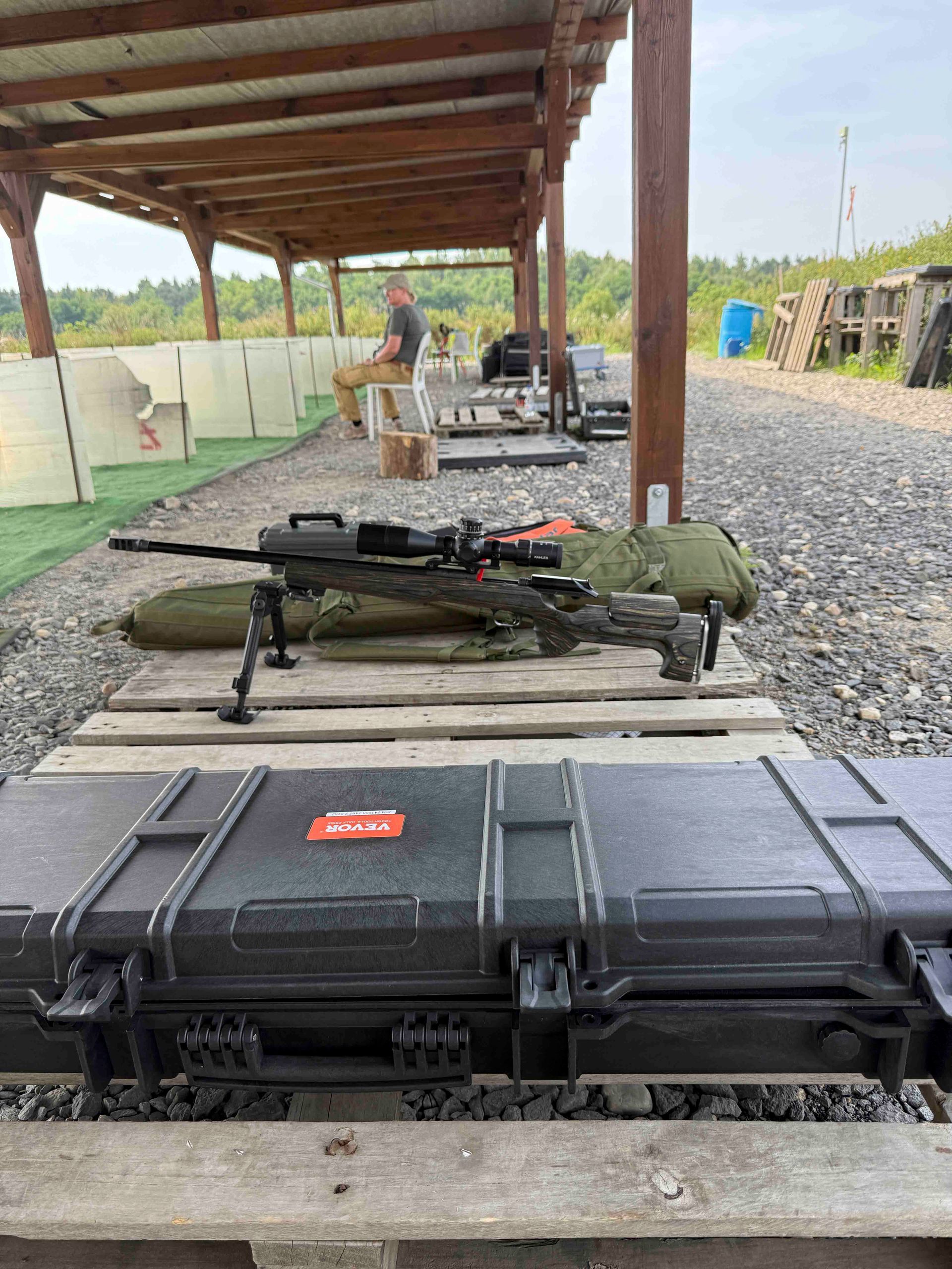 Ein Gewehr steht auf einer Holzpalette auf einem Schießstand im Freien. Im Hintergrund sitzt eine Person.