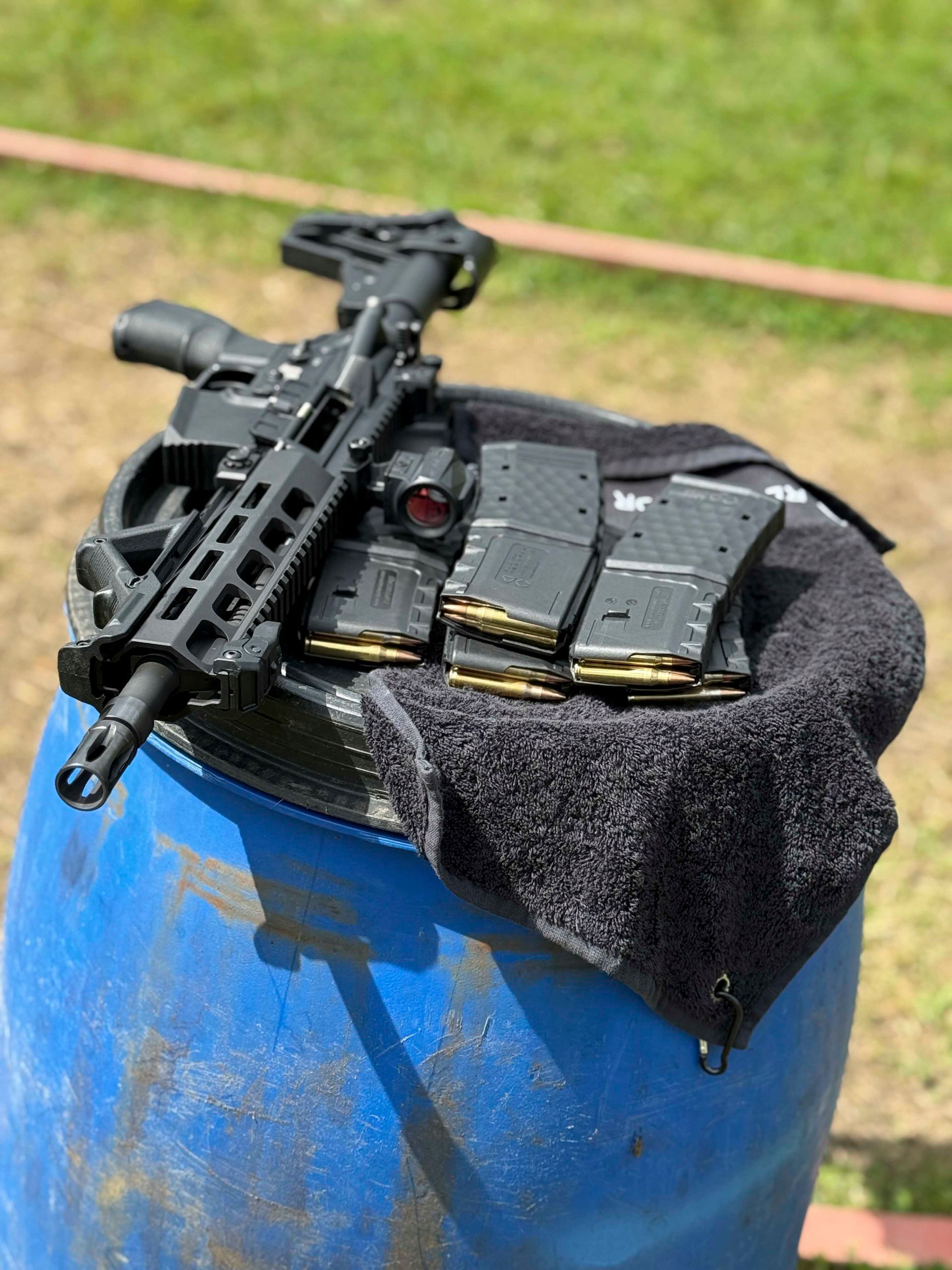 Schwarzes Gewehr, Magazine und Rotpunktvisier auf einem blauen Lauf, daneben ein Handtuch, draußen auf einer Wiese.