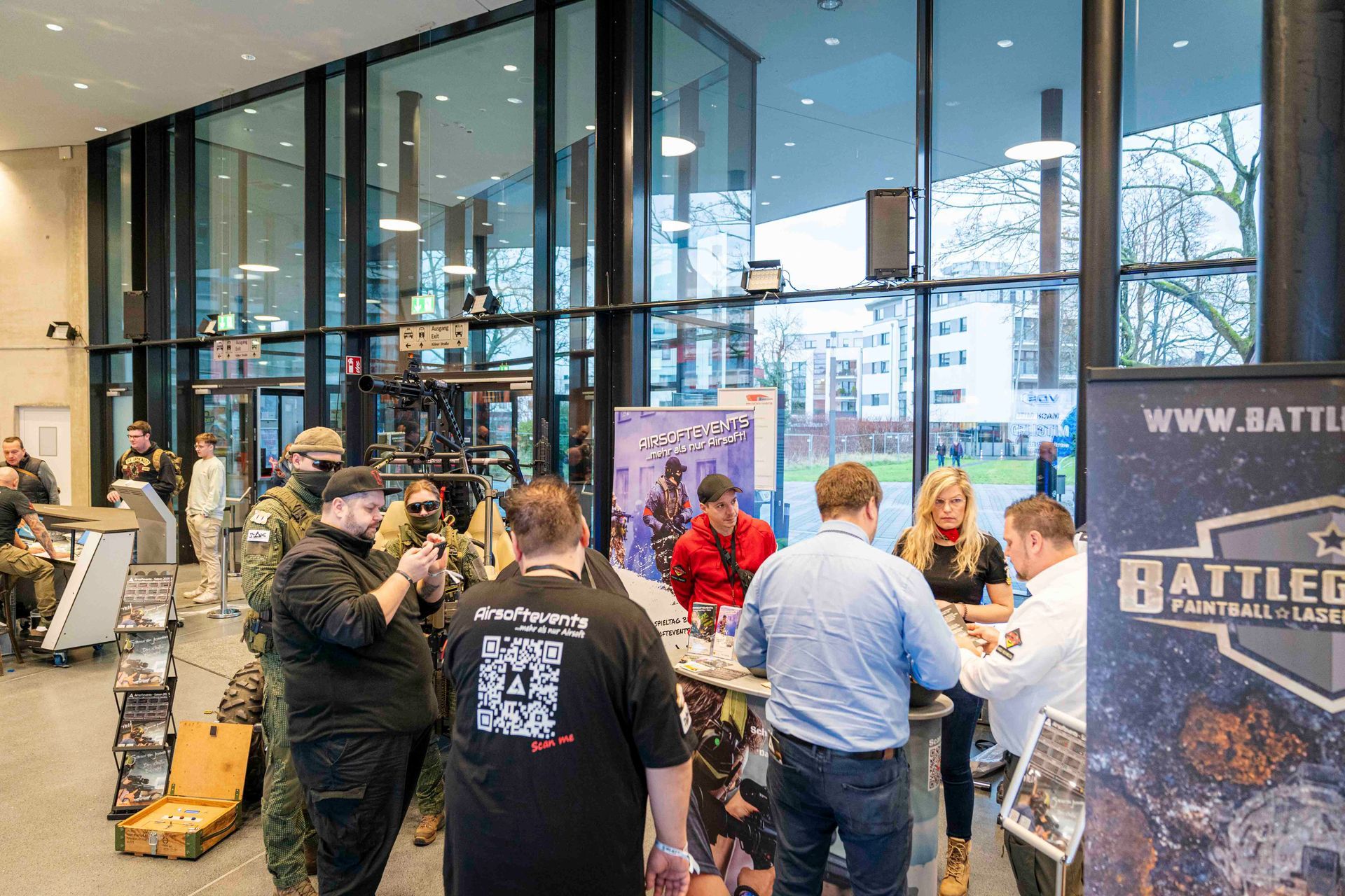 Menschen an einem Messestand, im Hintergrund große Fenster. Schild mit der Aufschrift „Battleground Paintball & Laser“.