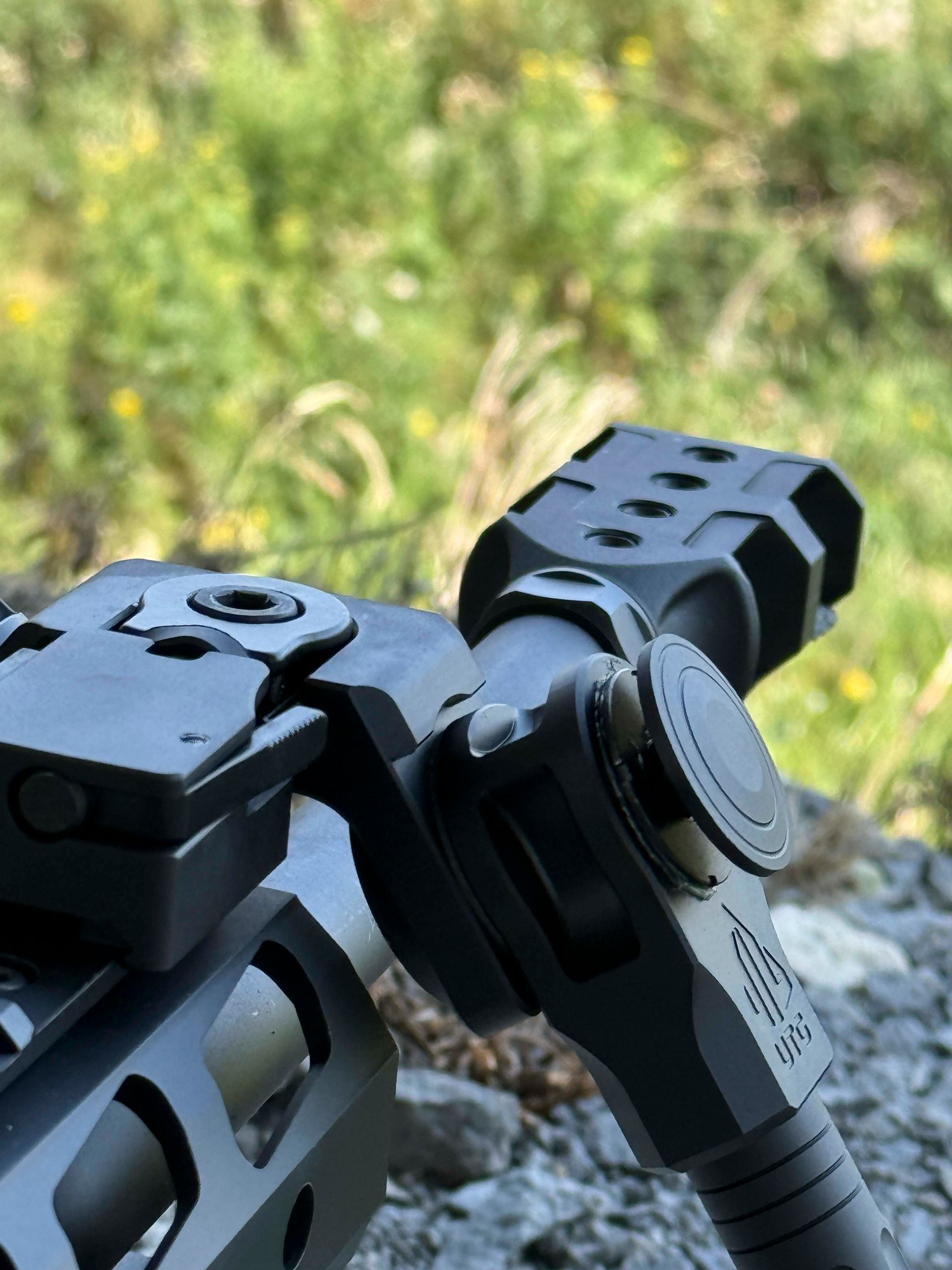 Schwarzes Zweibein für Gewehre im Fokus, im Freien vor verschwommenem grünem Laub.