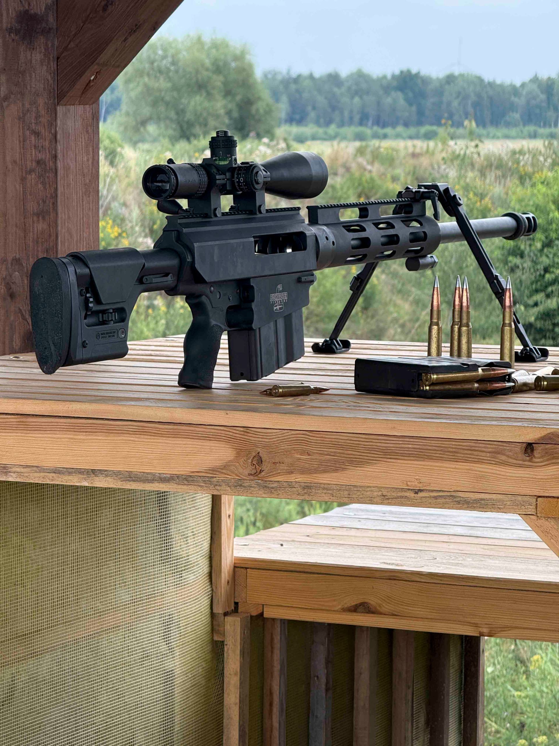 Schwarzes Scharfschützengewehr mit Zielfernrohr und Zweibein auf einem Holztisch, im Vordergrund liegen Patronen.