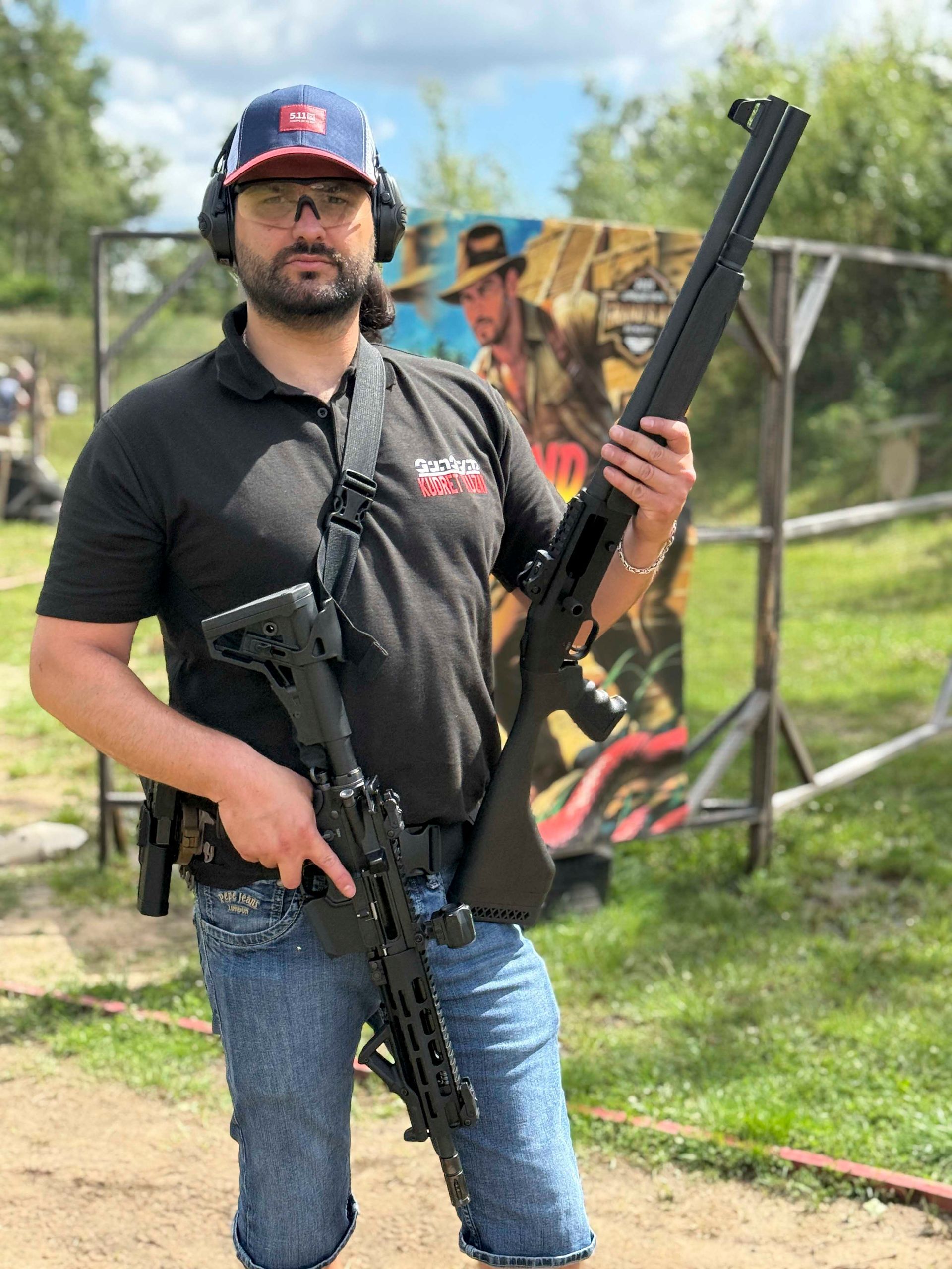 Ein Mann, der auf einem Schießstand im Freien eine Schrotflinte und ein Gewehr hält, trägt eine Baseballkappe und Gehörschutz