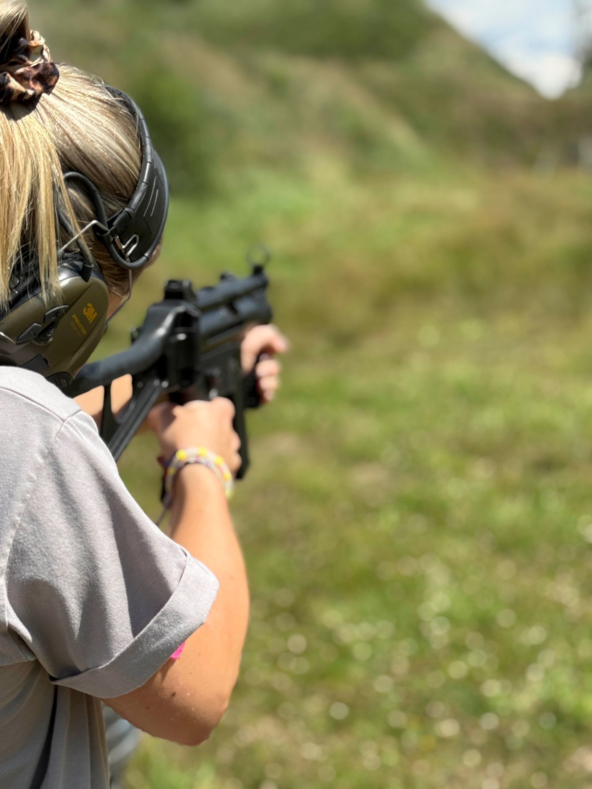 Frau zielt im Freien mit einer schwarzen Pistole; sie trägt Gehörschutz und ein helles Hemd; grüner Hintergrund.