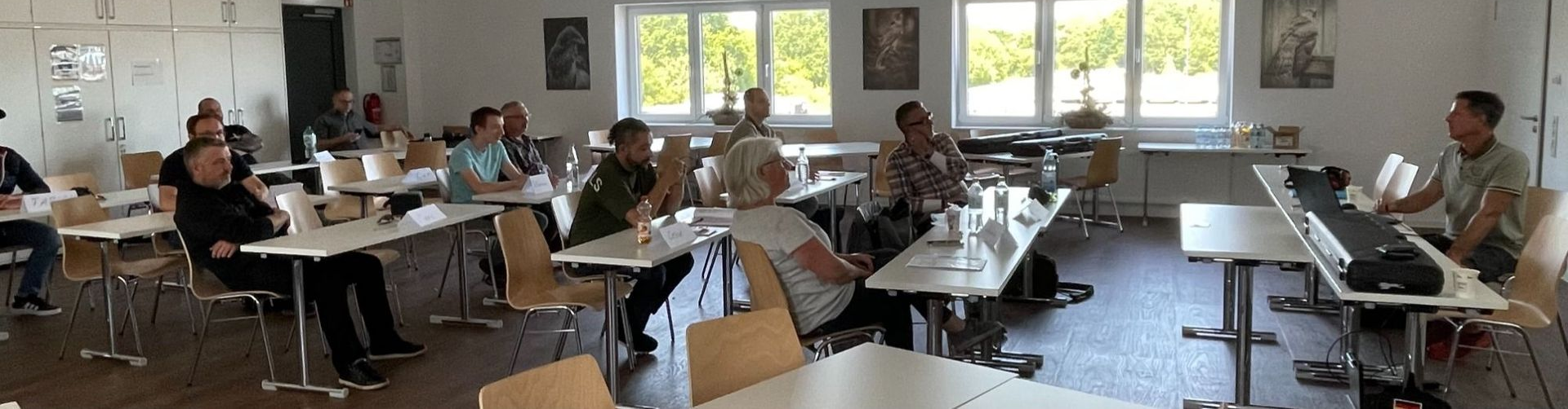 Menschen sitzen an Schreibtischen in einem hellen Klassenzimmer, einige benutzen während einer Gruppensitzung Laptops.
