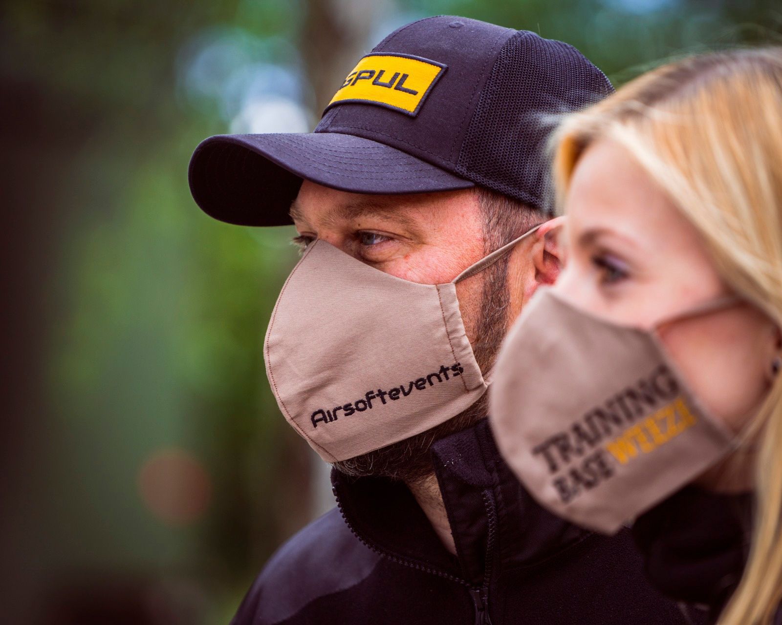 Ein Mann und eine Frau tragen Gesichtsmasken mit Veranstaltungslogos. Der Mann trägt eine Kappe.