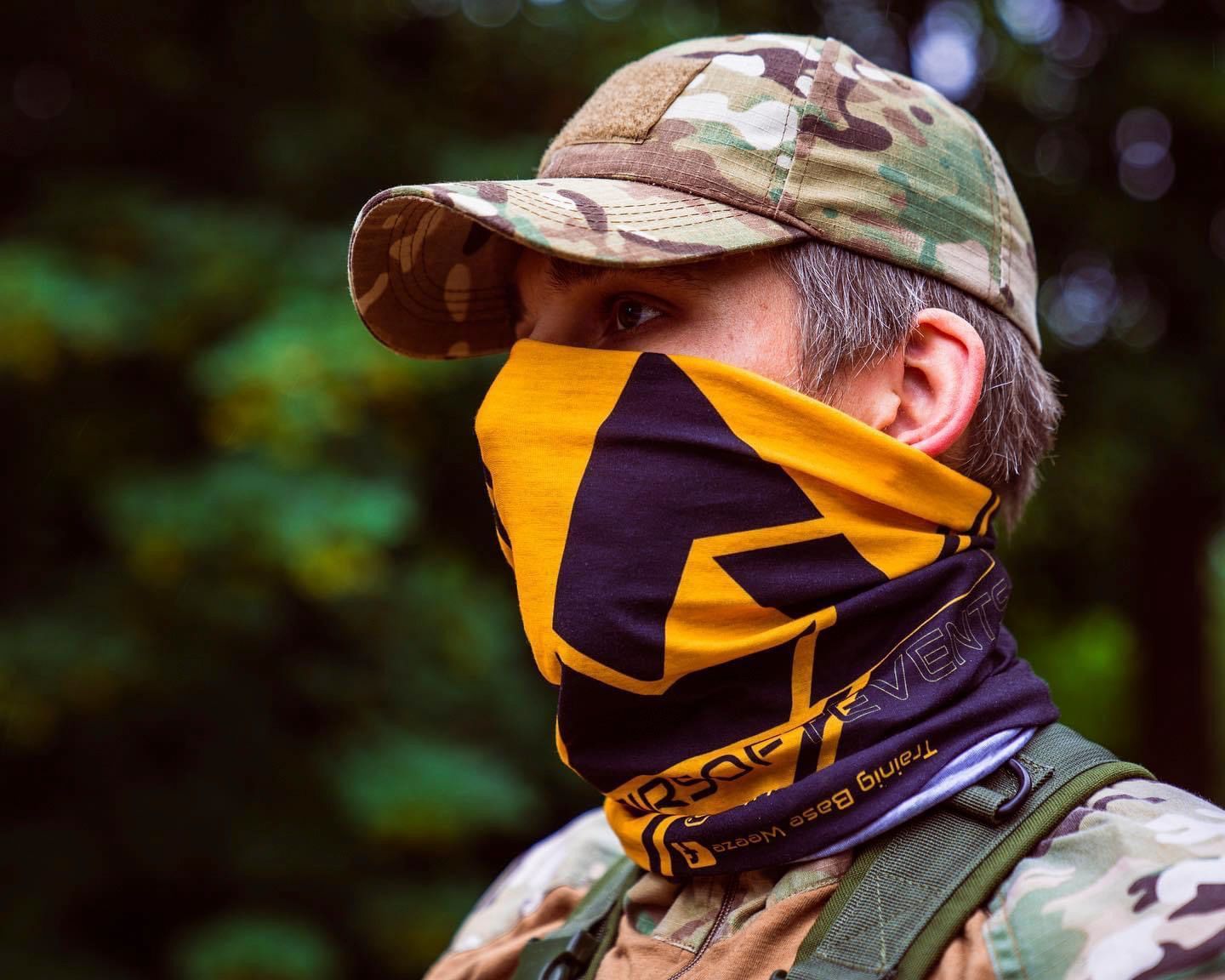 Person im Freien, die eine Tarnkappe und einen Halswärmer mit gelb-schwarzem Logo trägt.