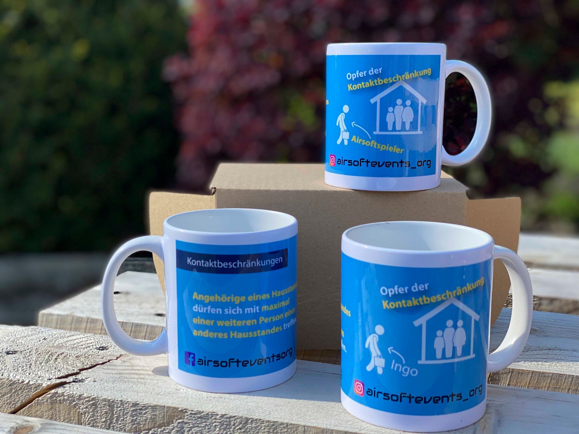 Drei weiße Becher mit blauer Schrift und Logos stehen draußen auf Holz.