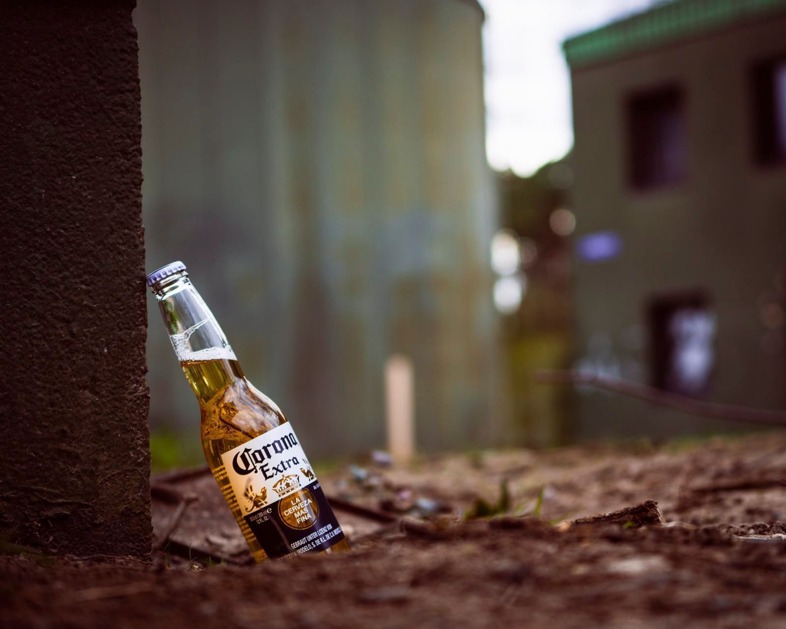 Eine Flasche Corona-Bier lehnt draußen an einem dunklen Pfosten auf braunem Boden, im Hintergrund sind Gebäude zu sehen.