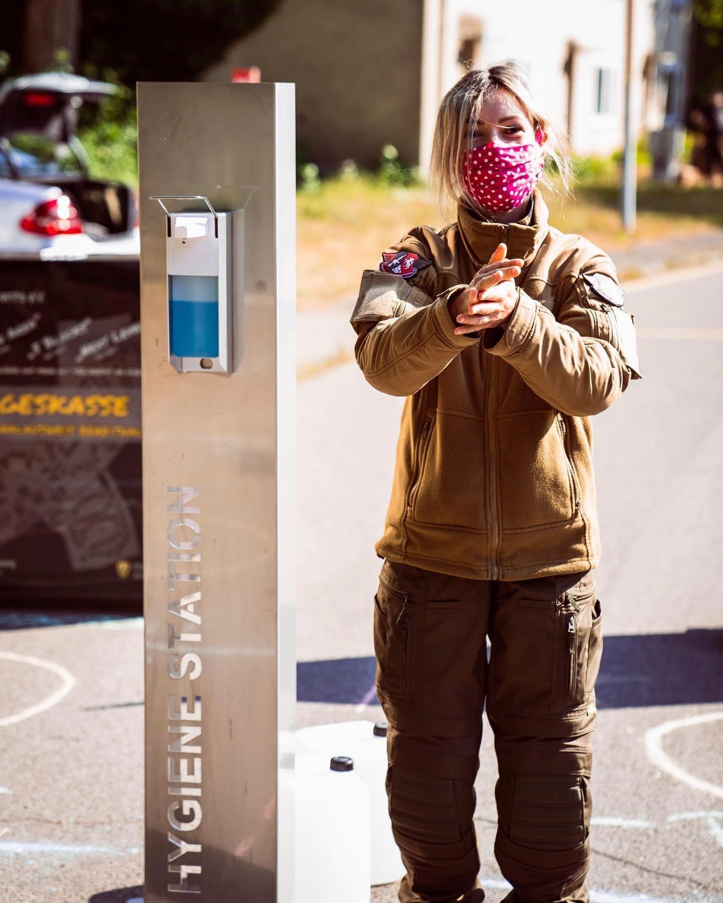 Frau benutzt Händedesinfektionsmittel an einer Hygienestation; trägt eine Maske und braune Kleidung im Freien.