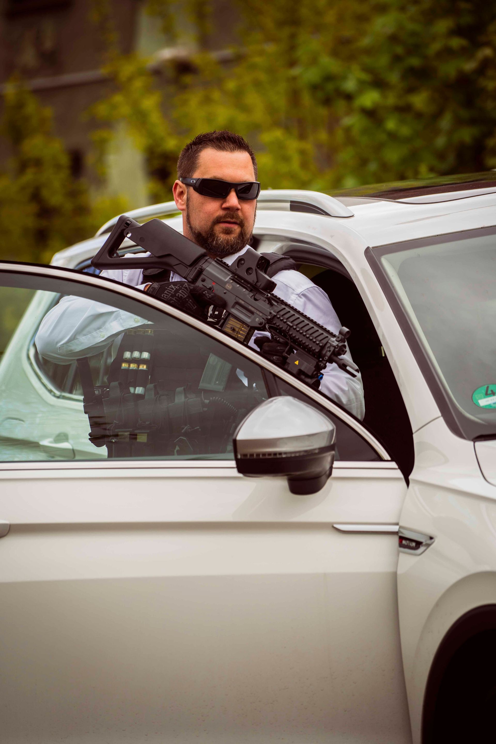 Ein Mann mit einem Gewehr lehnt aus einem silbernen Auto. Er trägt eine Sonnenbrille, sieht ernst aus, und die Tür ist offen.