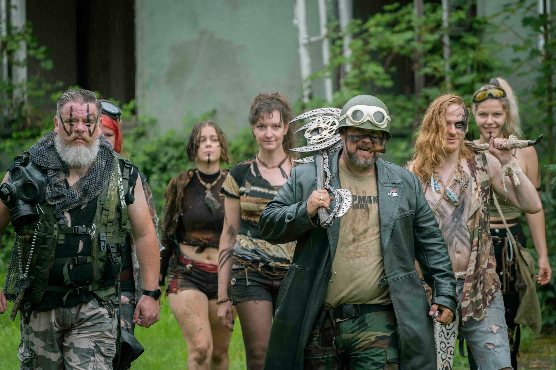 Eine Gruppe von Menschen in postapokalyptischer Kleidung wandert durch wucherndes Laubwerk 