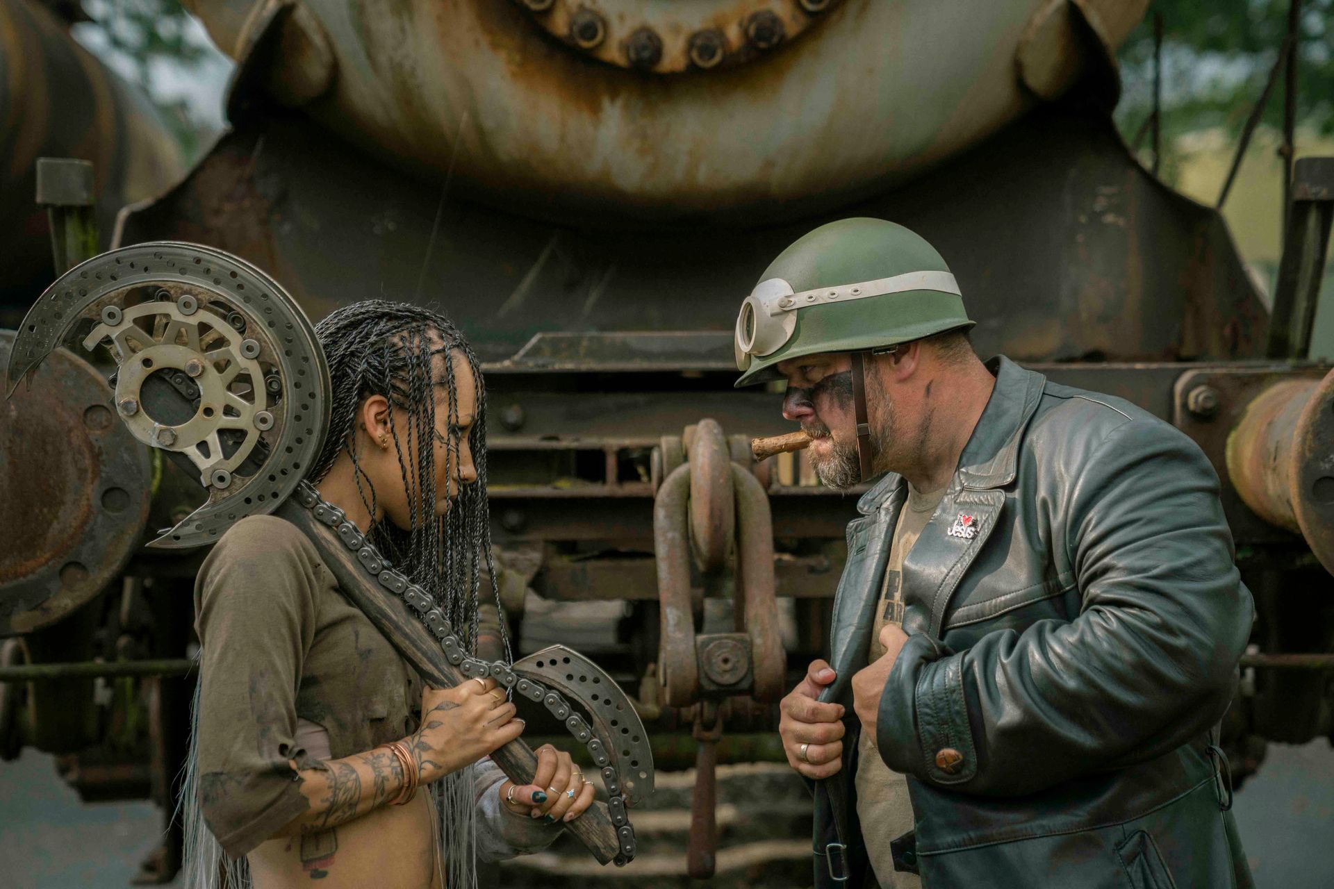 Frau und Mann in einer postapokalyptischen Welt. Sie hält eine Kette, er raucht. Im Hintergrund ein verwitterter Zug.