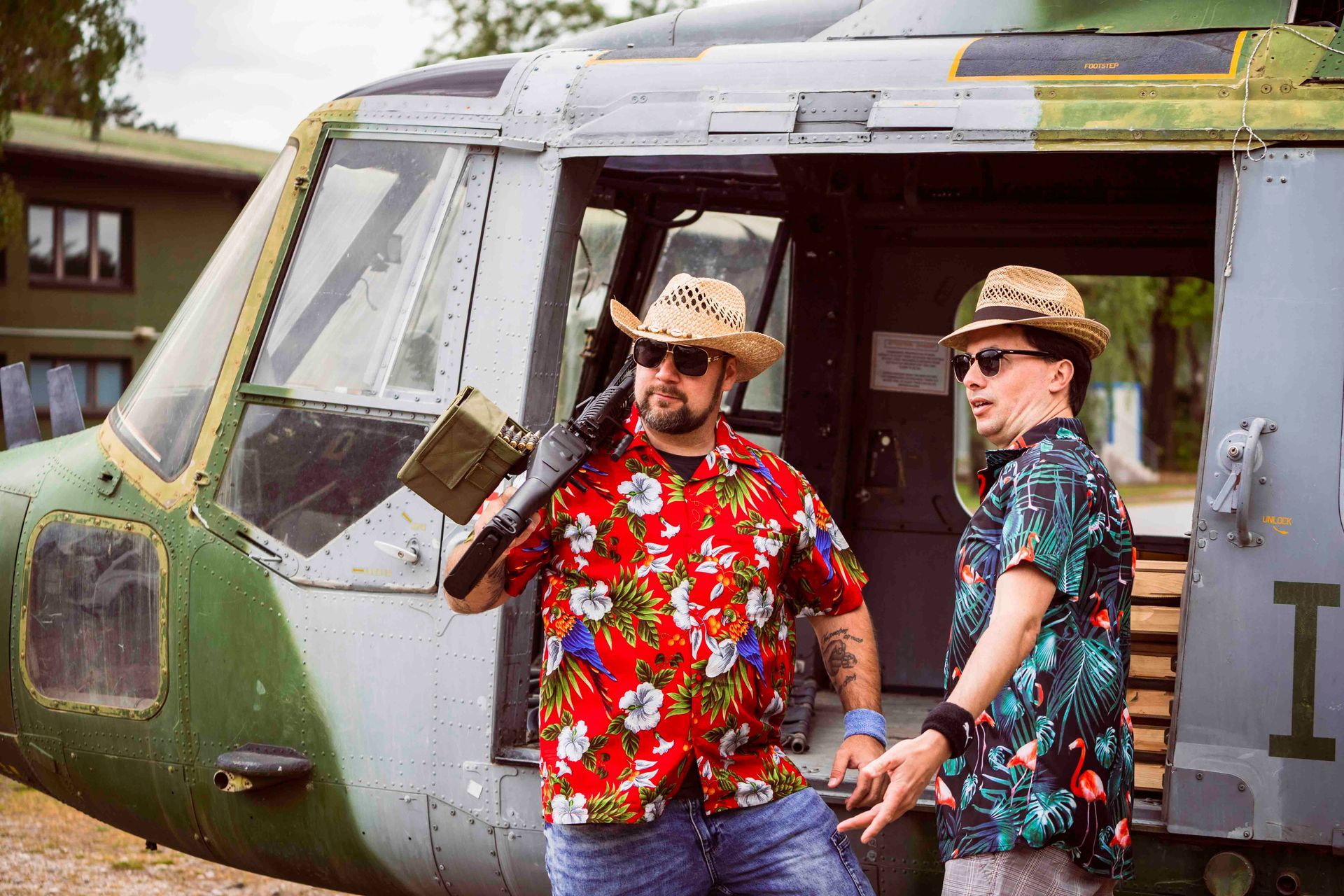 Zwei Männer mit Strohhüten und Hawaiihemden posieren neben einem Militärhubschrauber; einer hält eine Waffe.