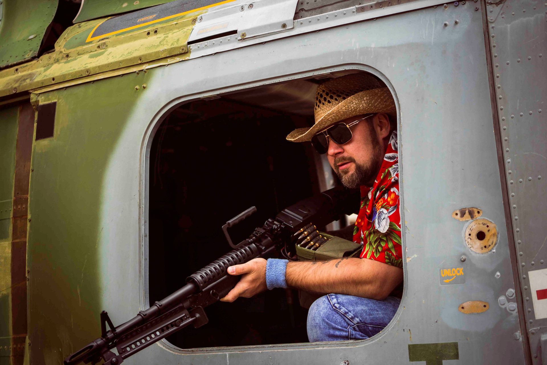 Mann mit Strohhut und Sonnenbrille, der ein Maschinengewehr hält und aus dem Fenster eines Hubschraubers schaut.