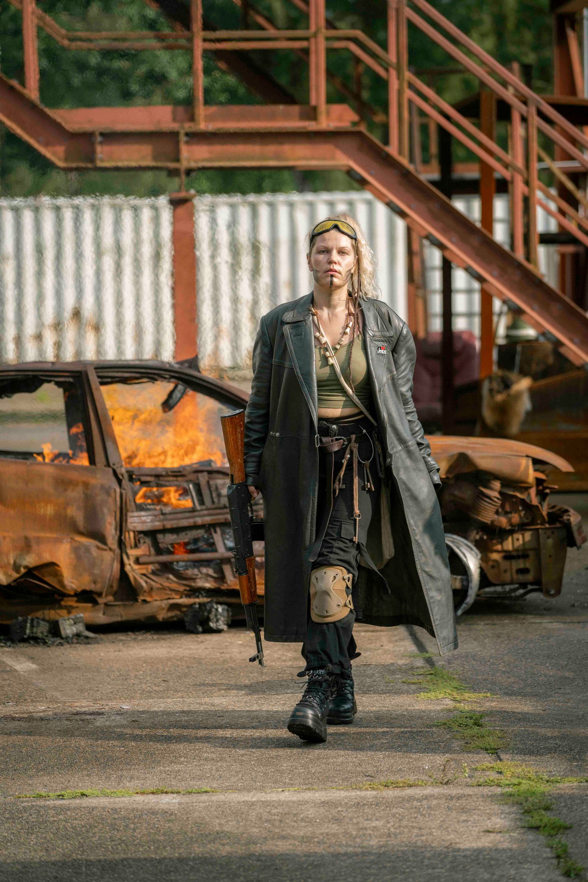 Eine Frau in postapokalyptischer Kleidung geht mit einem Gewehr in der Hand an einem zerstörten Auto vorbei. 