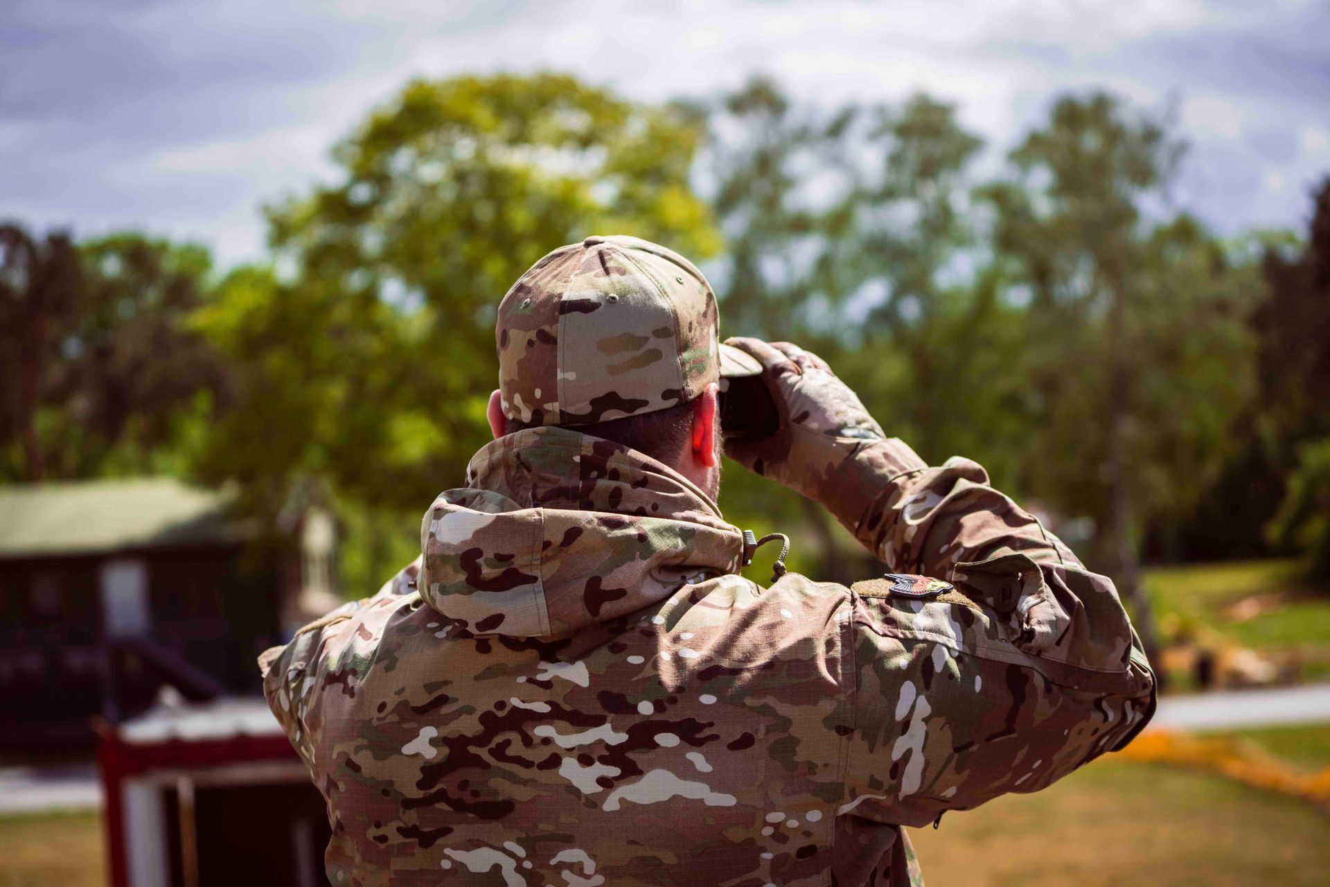 Ein Soldat in Tarnkleidung blickt im Freien durch ein Fernglas; im Hintergrund sind Bäume und Himmel zu sehen.
