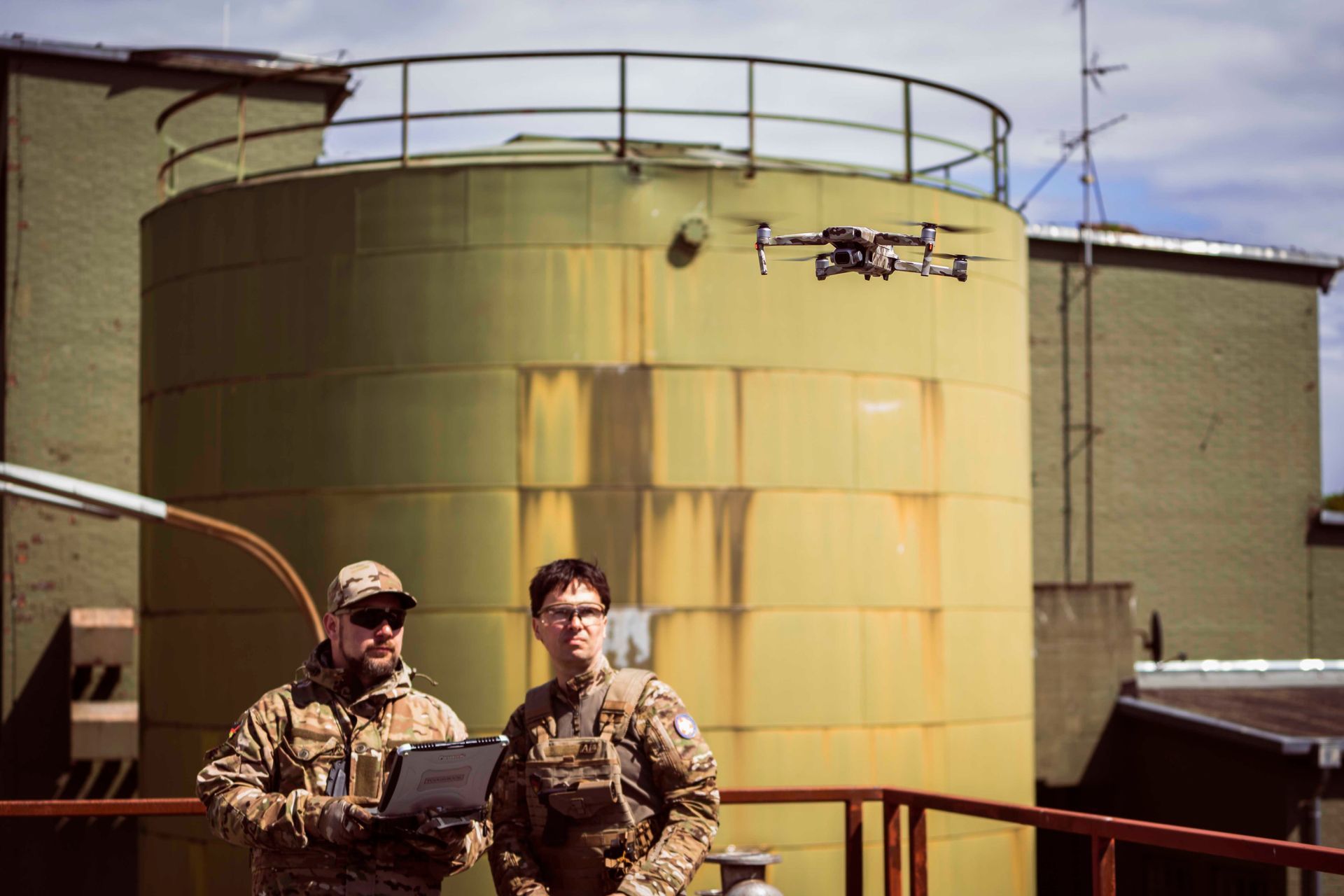 Zwei Personen in Militärkleidung bedienen eine Drohne in der Nähe eines großen zylindrischen Tanks auf einem Dach.