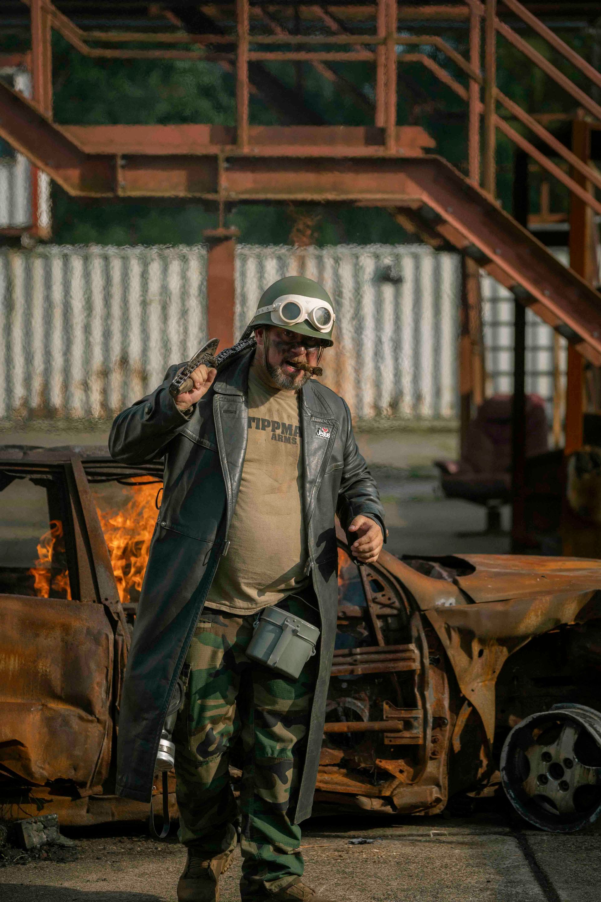 Ein Mann in postapokalyptischer Kleidung hält einen Schraubenschlüssel neben einem brennenden Auto.