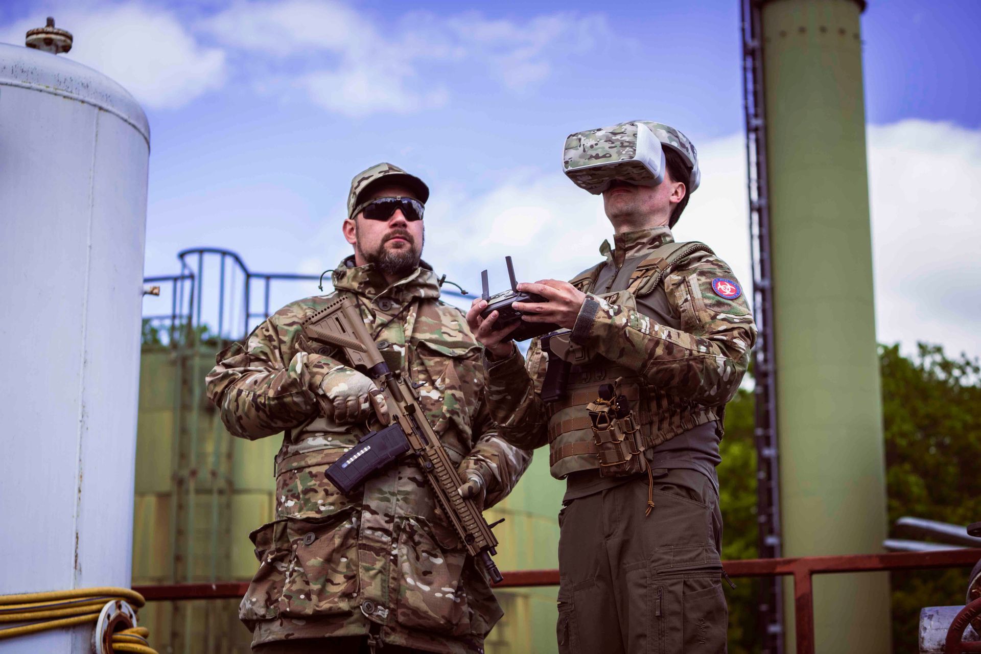 Zwei Personen in Tarnkleidung auf einem Dach; eine benutzt ein VR-Headset und eine Fernbedienung, die andere hält ein Gewehr.
