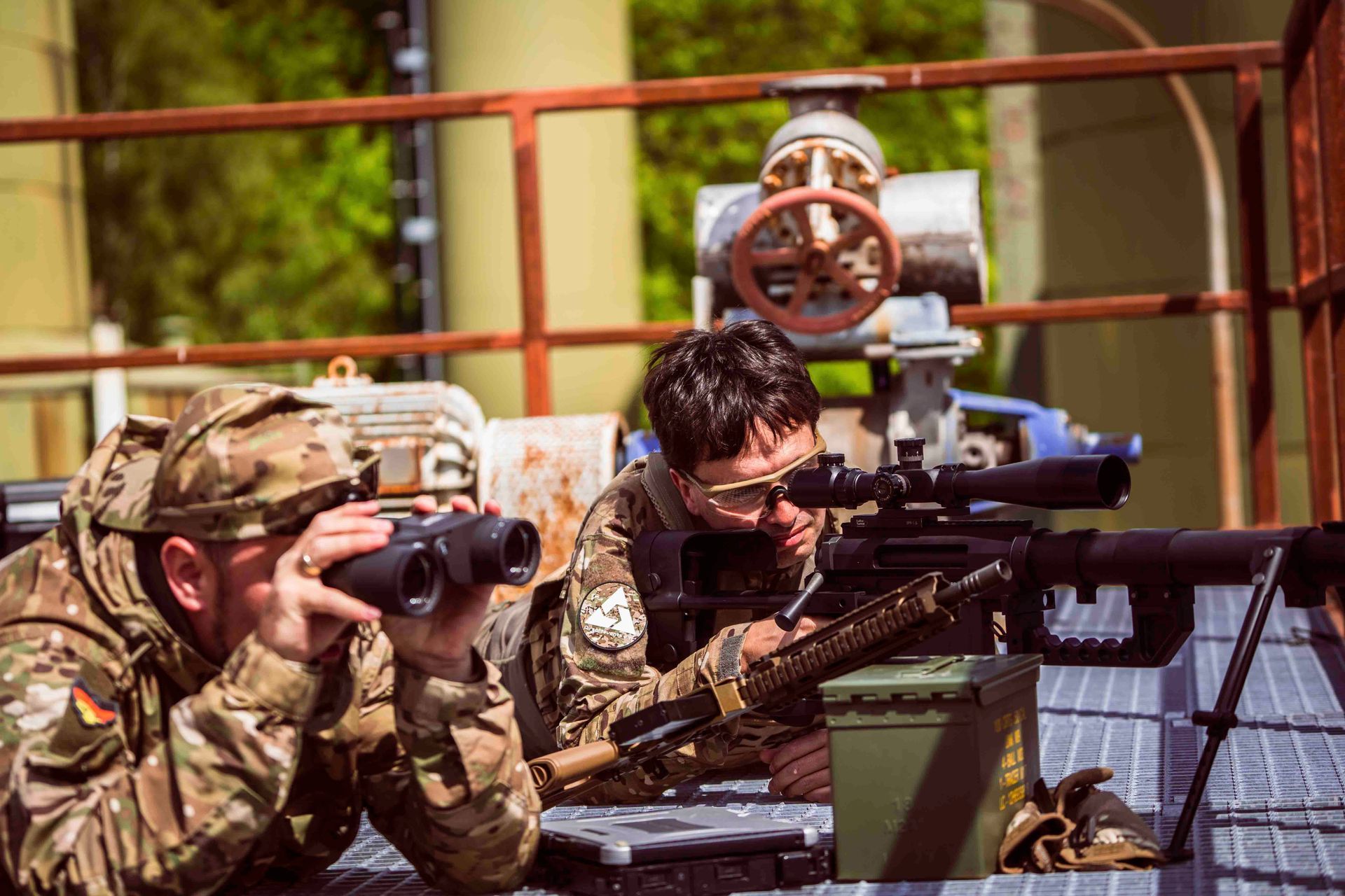 Zwei Soldaten in Tarnkleidung auf einer Plattform, einer benutzt ein Fernglas, der andere zielt mit einem Gewehr
