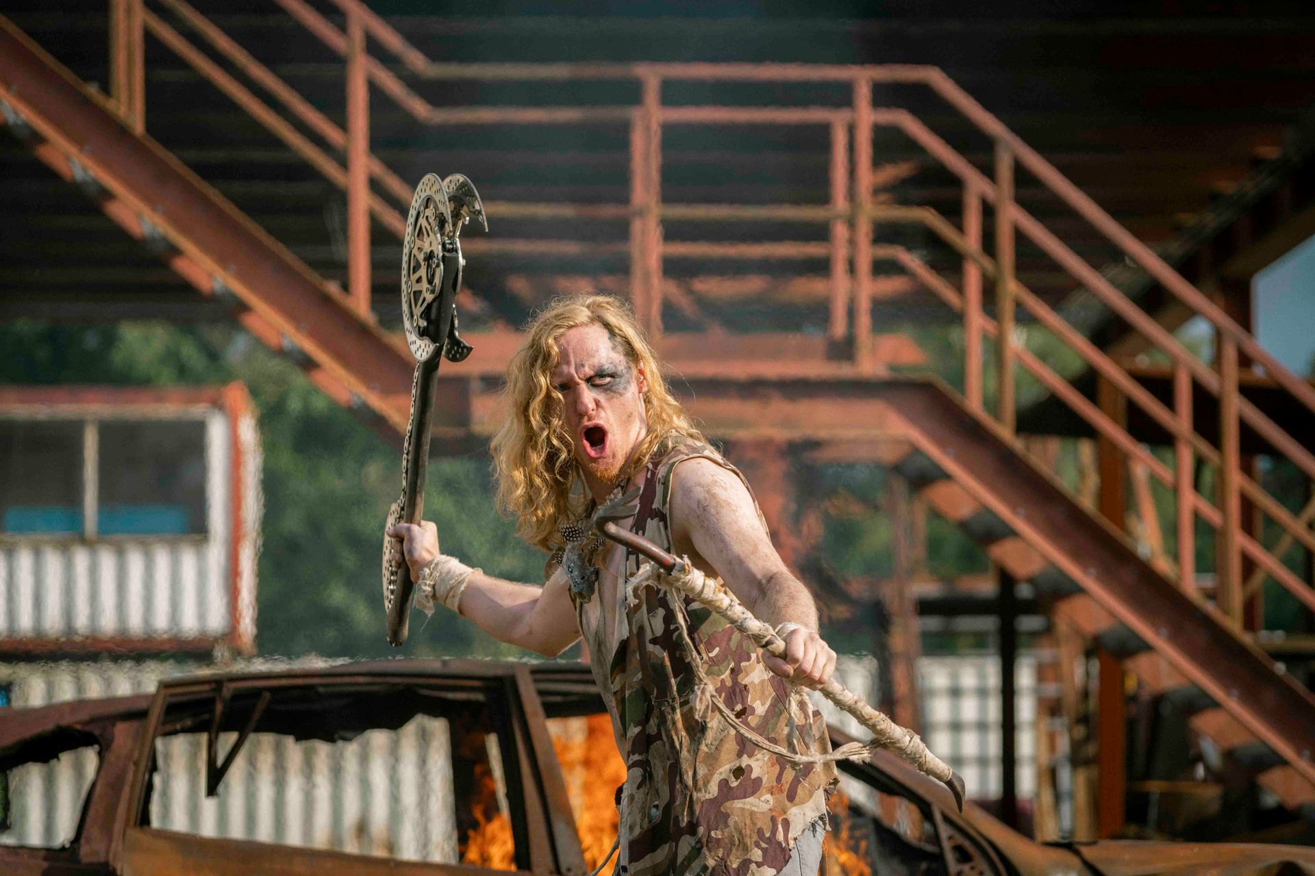 Eine Person mit langen blonden Haaren schreit und hält eine Waffe. Sie steht vor einem rostigen Gebäude