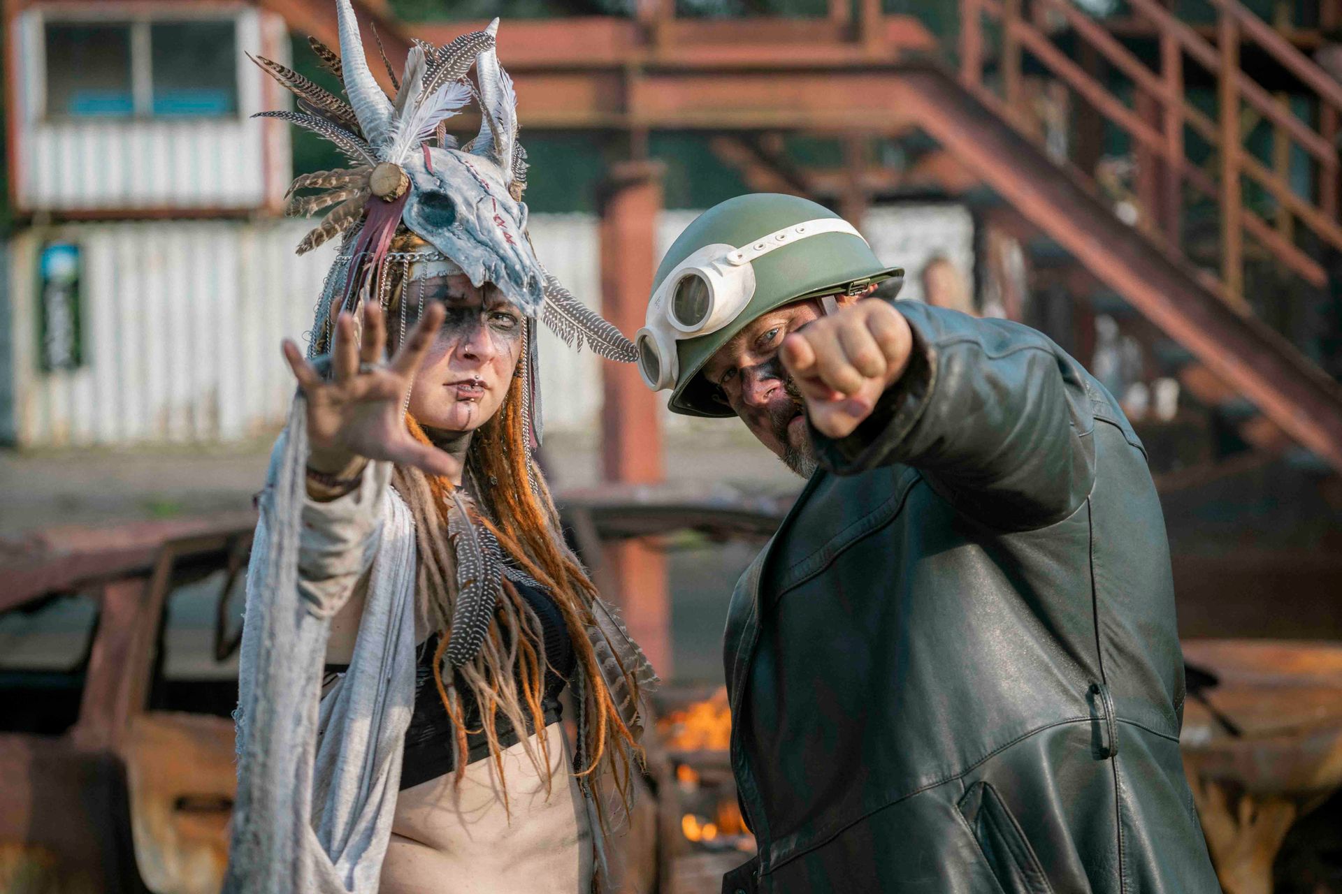 Zwei Personen posieren vor einem ausgebrannten Auto. Die eine trägt einen Kopfschmuck, die andere einen Helm