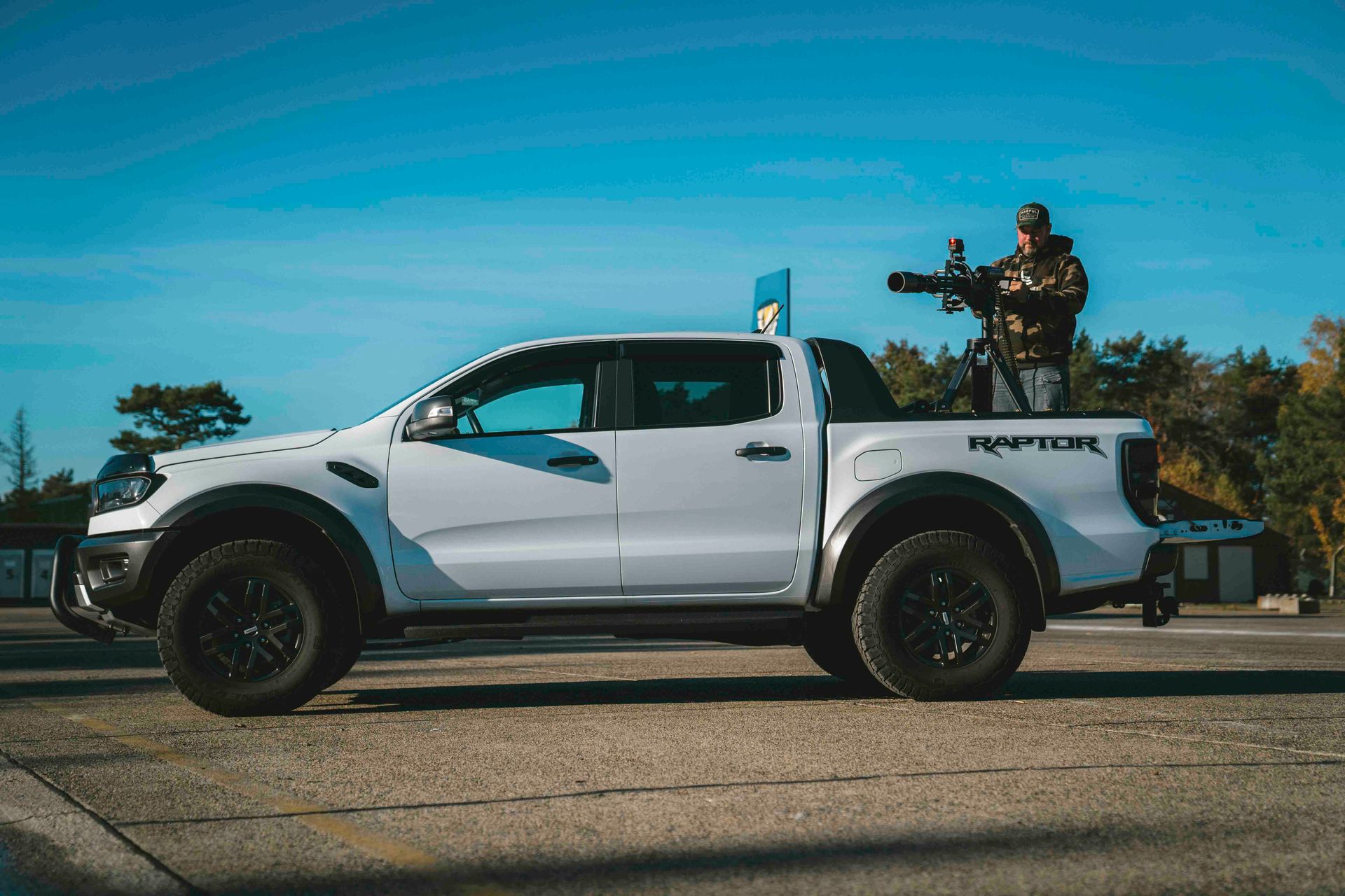 Weißer Ford Ranger Raptor Pickup mit einer Person, die auf der Ladefläche eine Kamera hält. Blauer Himmel im Hintergrund.