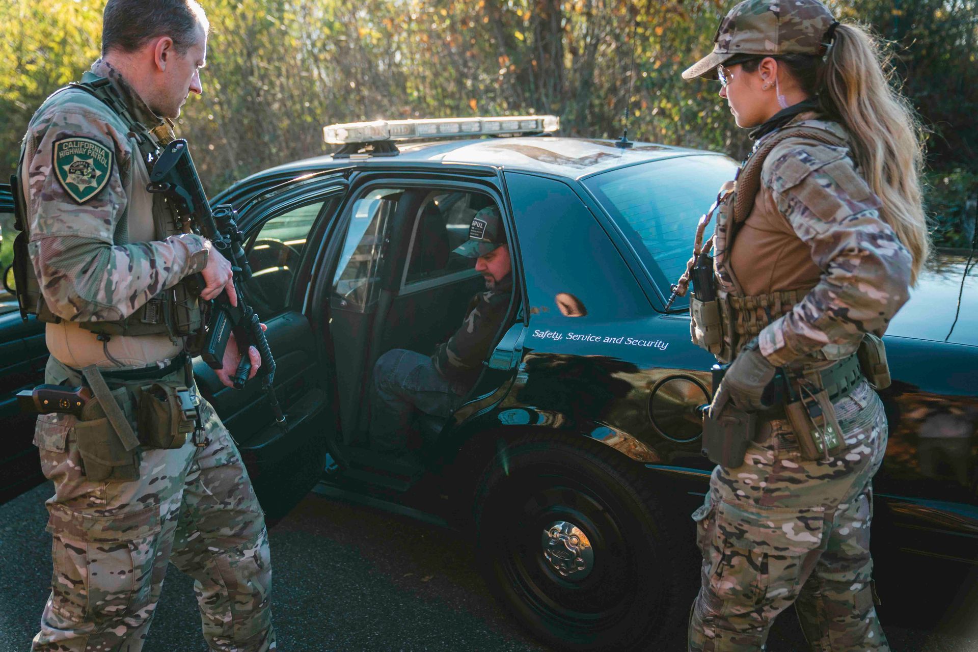 Zwei Polizeibeamte in Tarnkleidung flankieren einen Polizeiwagen; eine Person steigt aus dem Wagen aus.