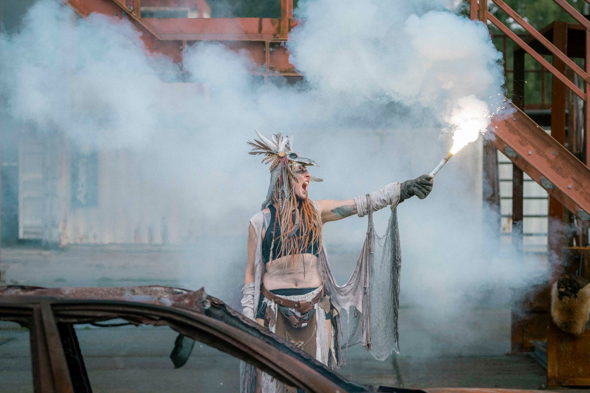 Eine Person in aufwendigem Kostüm hält eine Leuchtrakete und steht inmitten von Rauch und Trümmern.