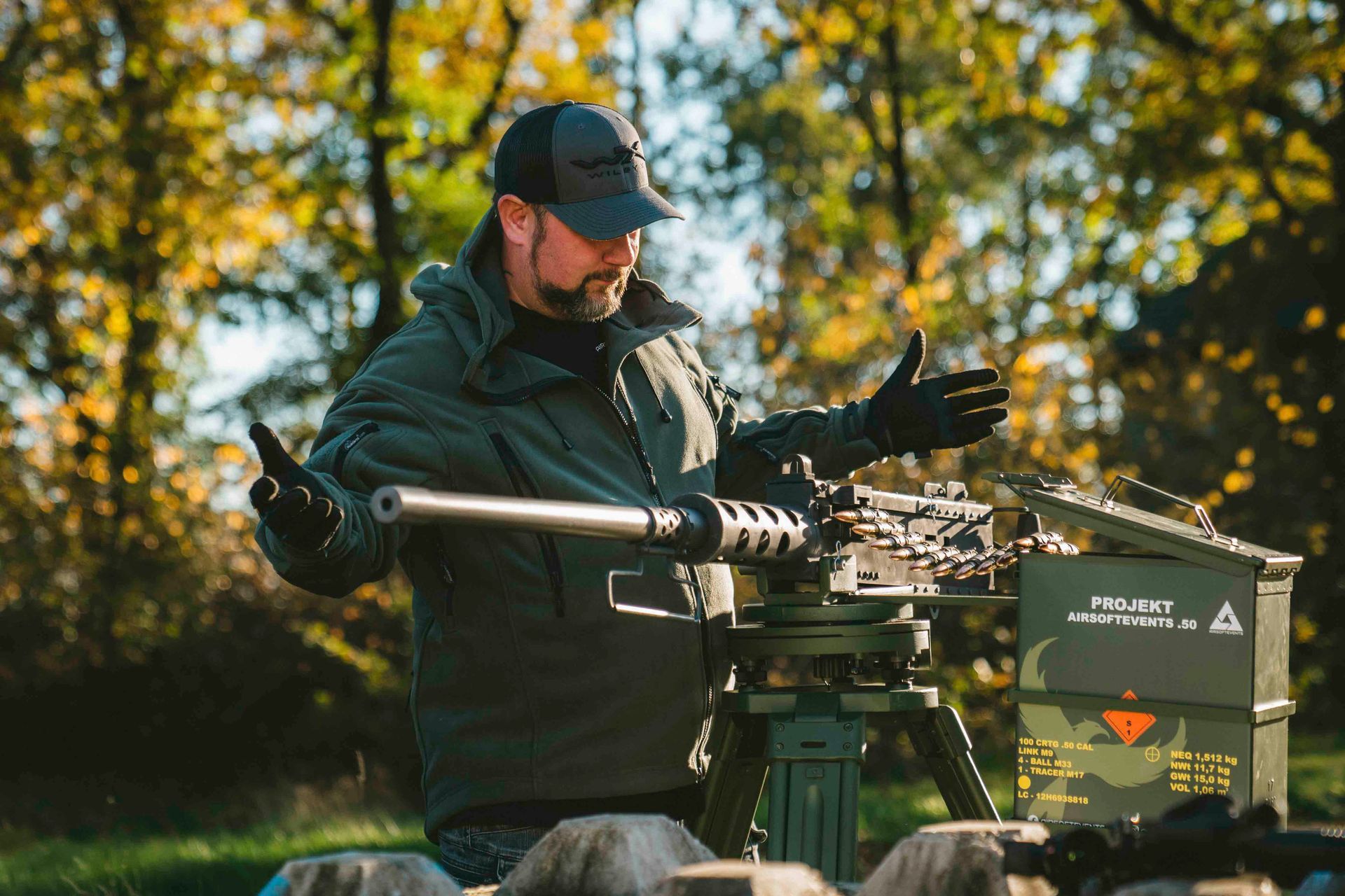 Mann mit Hut und Handschuhen inspiziert im Freien ein montiertes Maschinengewehr; im Hintergrund sind Bäume zu sehen.