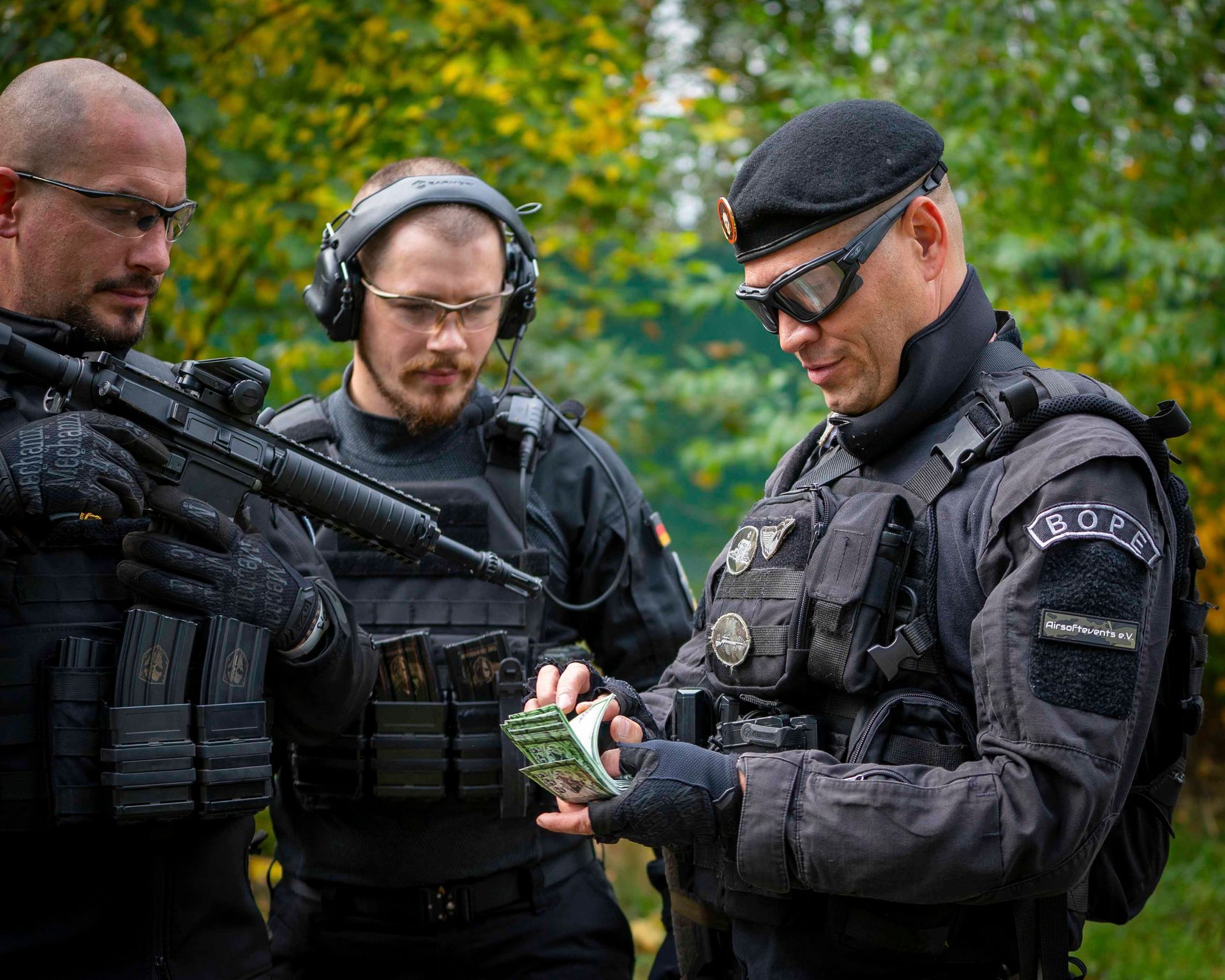Drei bewaffnete Männer in taktischer Ausrüstung untersuchen im Freien, im Waldhintergrund, einen Geldstapel.